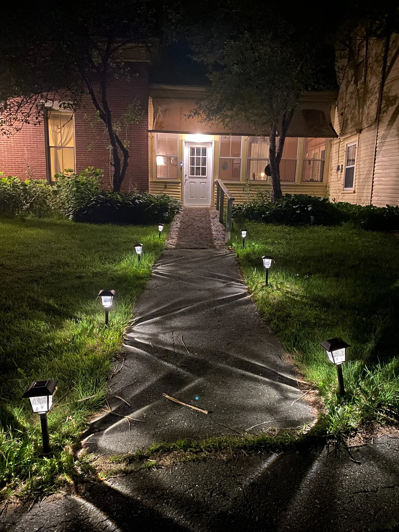 A house with a porch illuminated by lights at night, with a pathway lined with small ground lights, and trees casting shadows on the sidewalk.