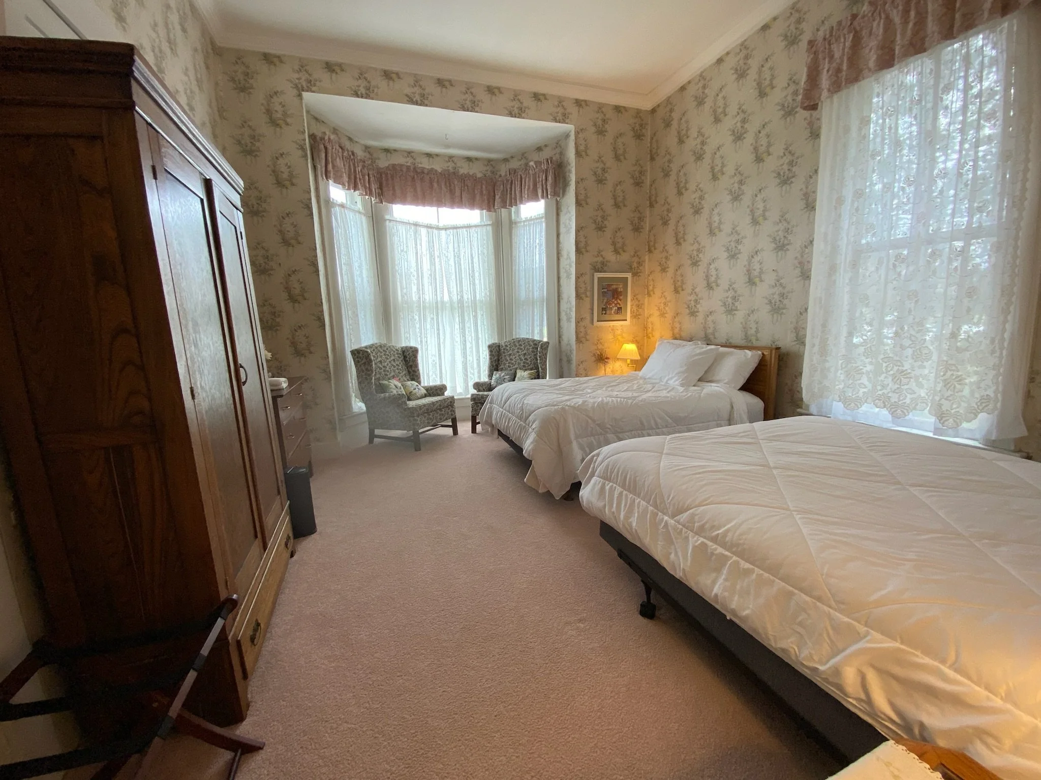 A bedroom with floral wallpaper, featuring two beds with white bedding and pillows, a wooden headboard, a window with sheer curtains, two framed black-and-white photographs on the wall, a nightstand with a lamp. Perfect for children or a small group.