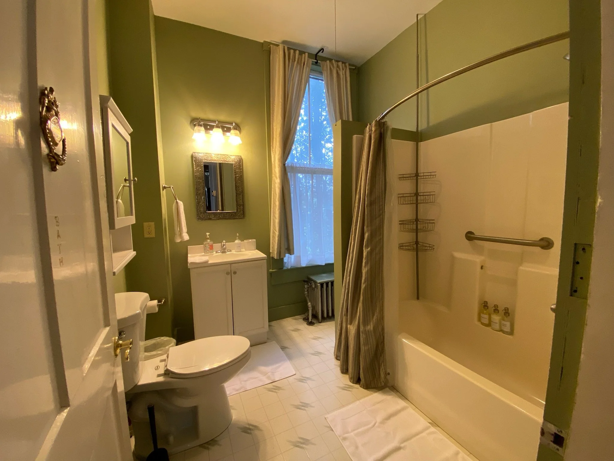 A small bathroom with a toilet, a vanity with a mirror, a shower stall, and a towel hanging on a hook. The floor is tiled with light-colored tiles, and there is a small red stool in front of the vanity.