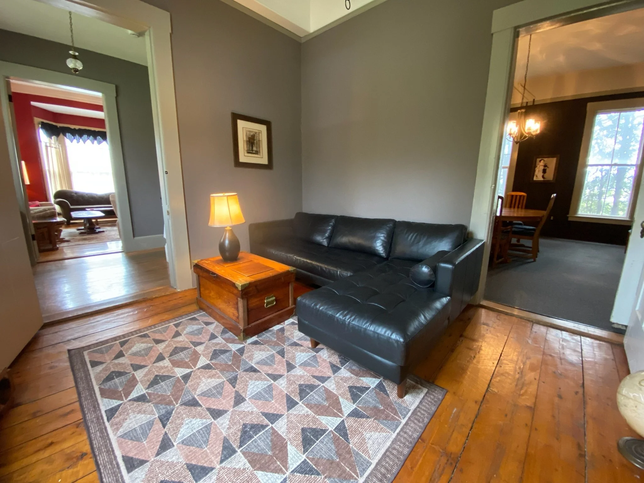 Living room with a black leather sofa, a wooden coffee table with a lamp, a patterned rug, and walls decorated with framed pictures. Adjacent rooms visible through doorways, including a dining area with a chandelier and another room with large windows and red walls.