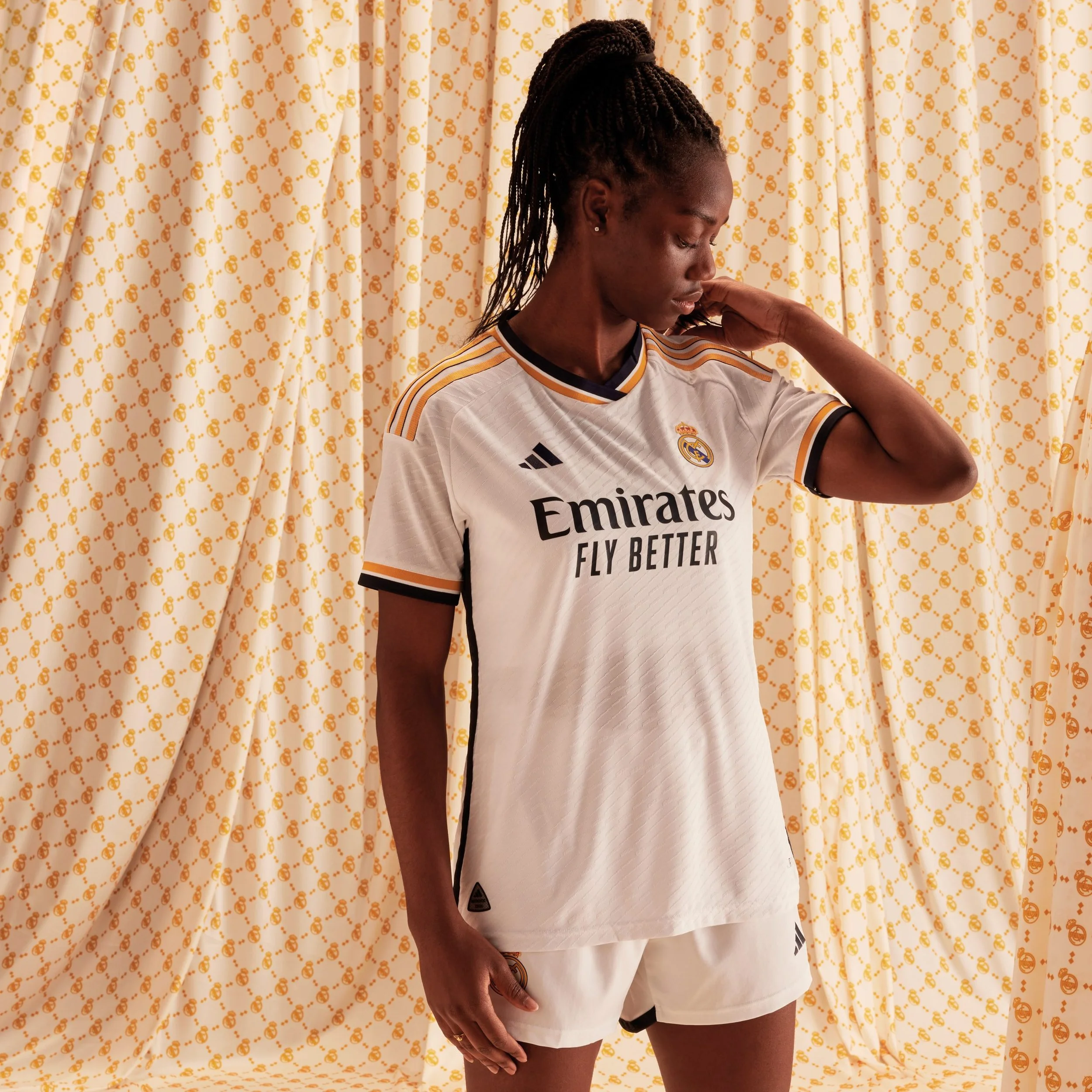 Una mujer con uniforme de fútbol de adidas Real Madrid frente a cortinas decorativas de color crema con patrones dorados. campaña AMOR INCONDICIONAL