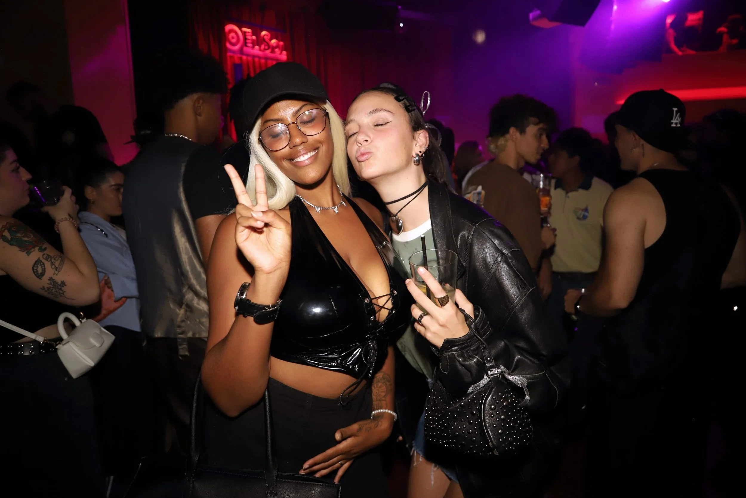 evento campaña LFSTLR adidas Real Madrid - Dos mujeres en una fiesta, una sonriendo y haciendo una señal de paz, ambas con ropa llamativa y accesorios, rodeadas de personas y luces de colores.