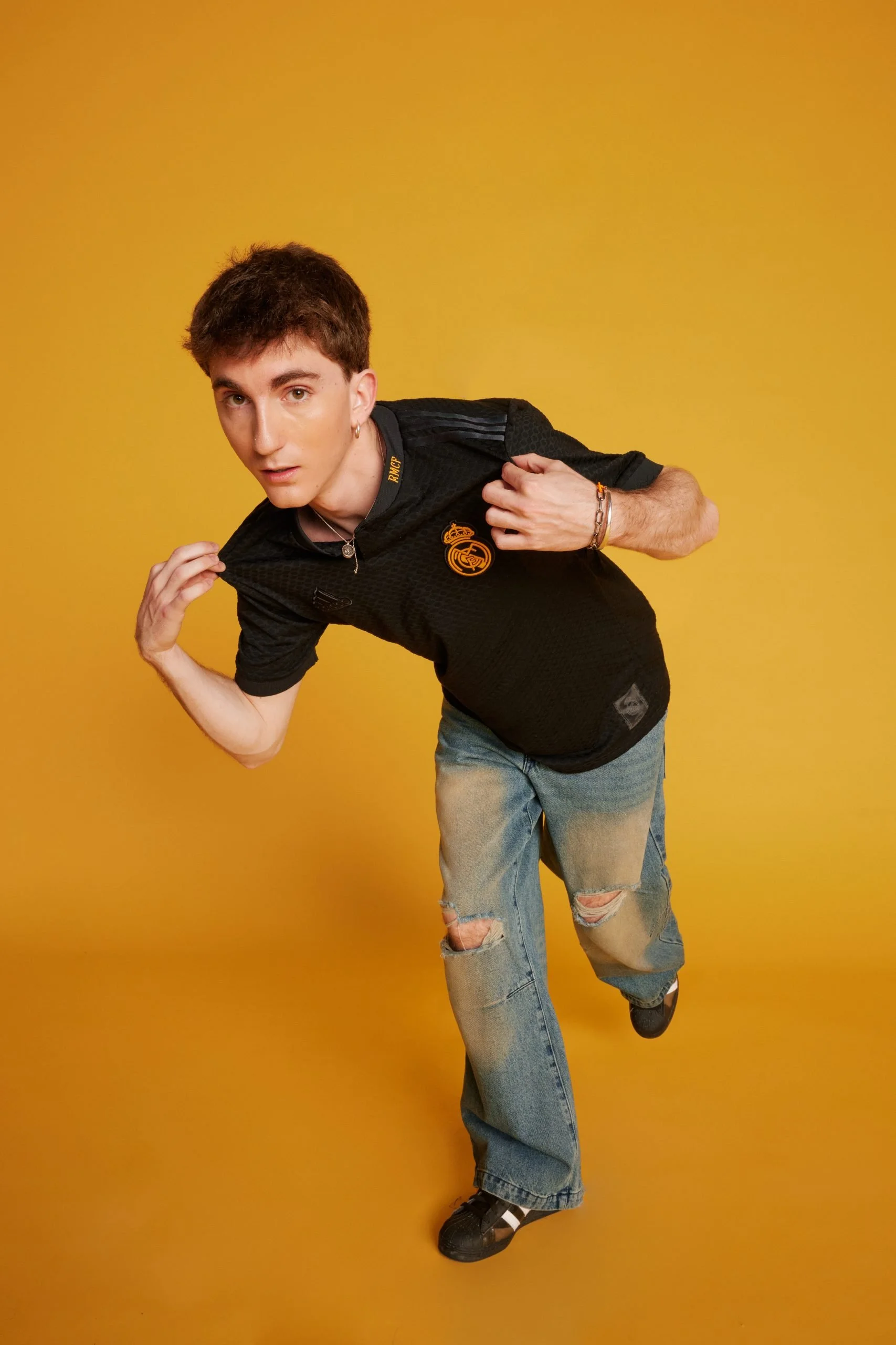 Joven vistiendo camiseta negra campaña LFSTLR adidas Real Madrid, pantalones vaqueros rotos, posando en un fondo amarillo.