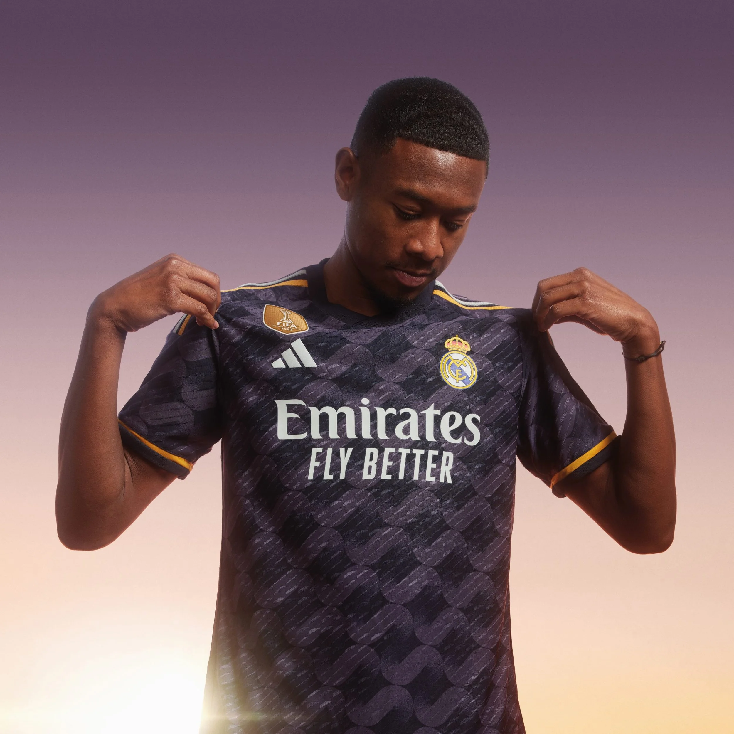 Joven hombre con piel oscura levantando el cuello de su camiseta de fútbol del Real Madrid, con logotipos y patrocinadores visibles, en un fondo de atardecer.