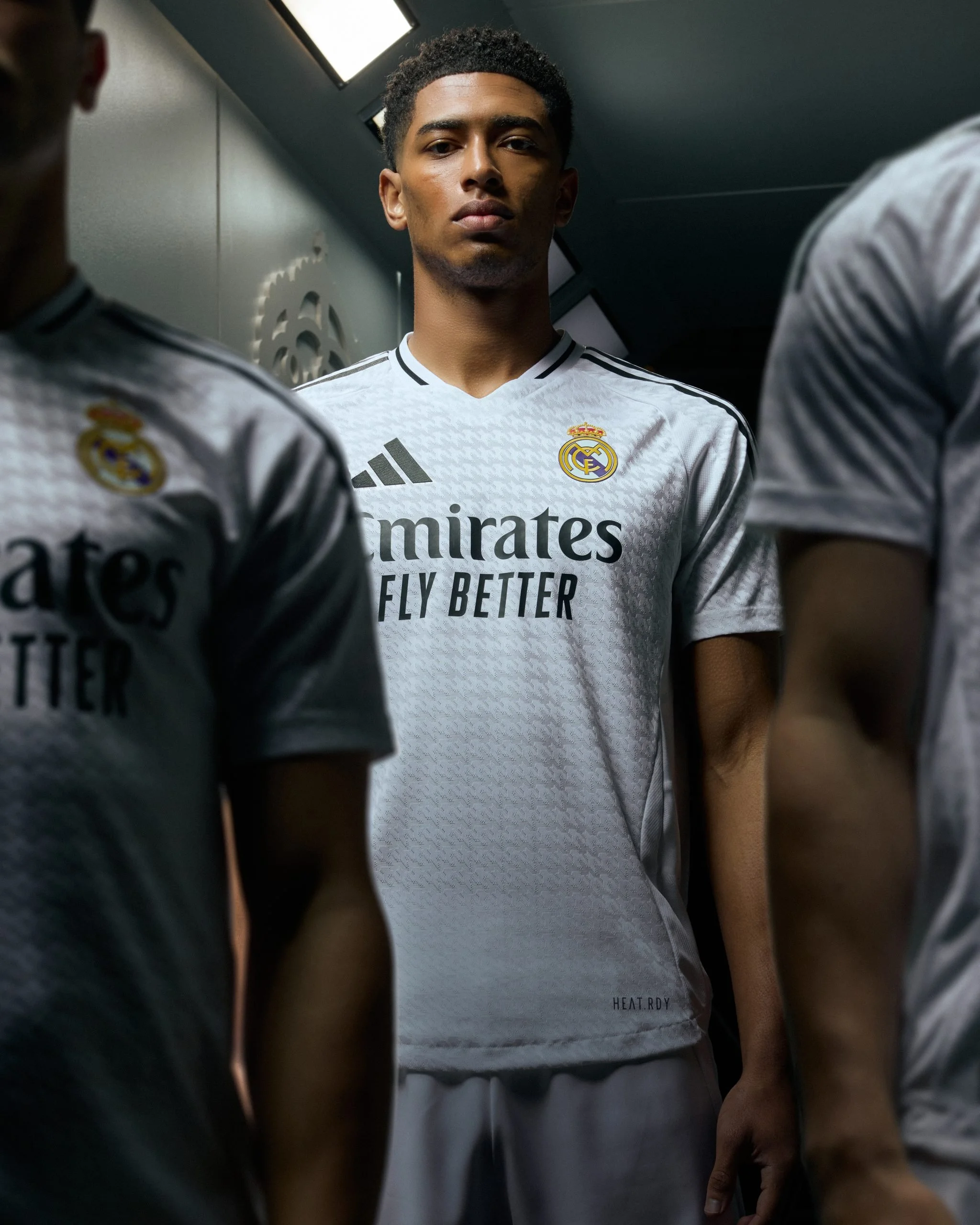 Joven futbolista de pie en un vestidor, vestido con uniforme de adidas Real Madrid, rodeado de otros jugadores. Él mira serio y concentrado.