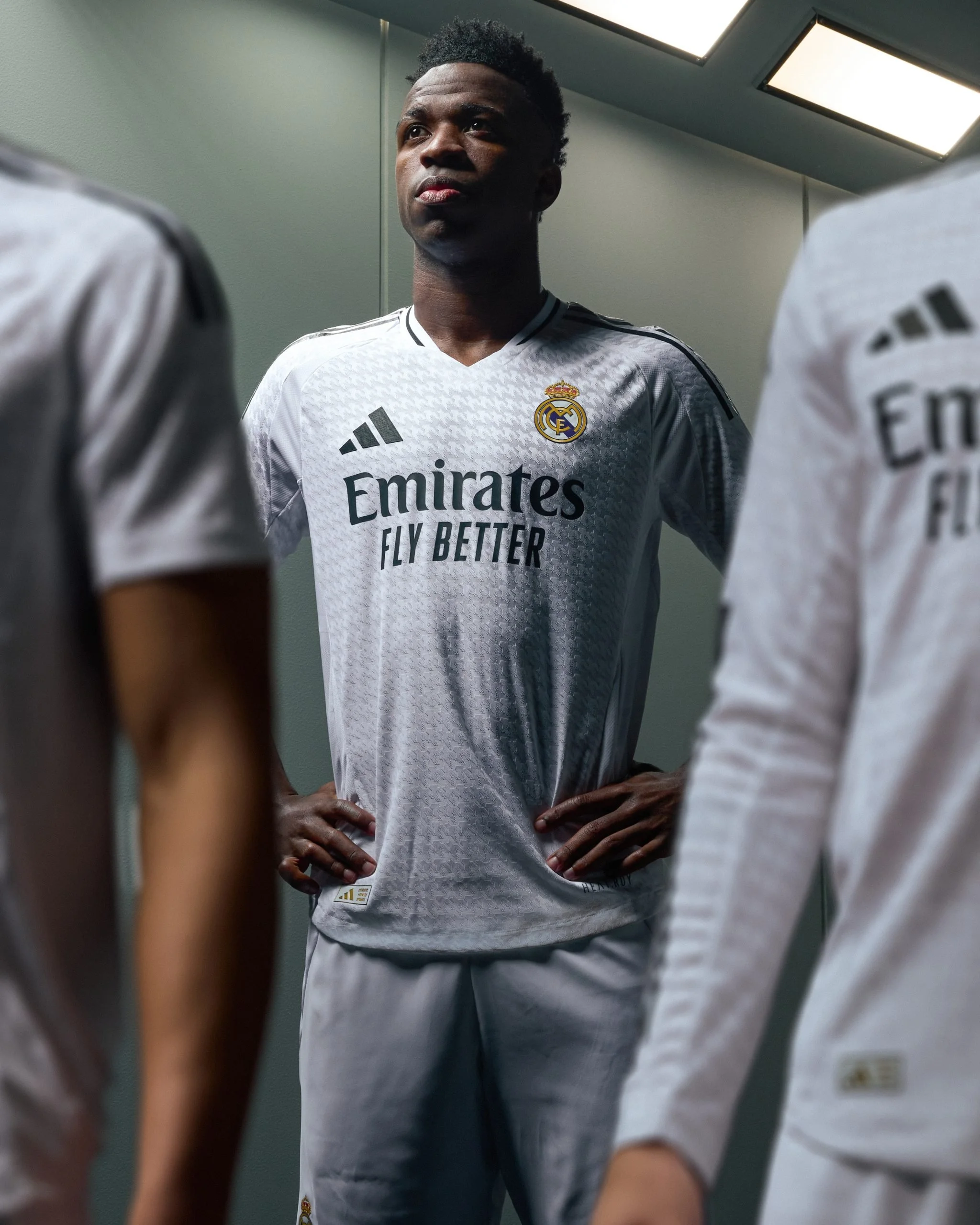 Joven futbolista en vestimenta adidas del Real Madrid, de pie con manos en las caderas, en un vestuario con iluminación moderna, rodeado de otros jugadores.