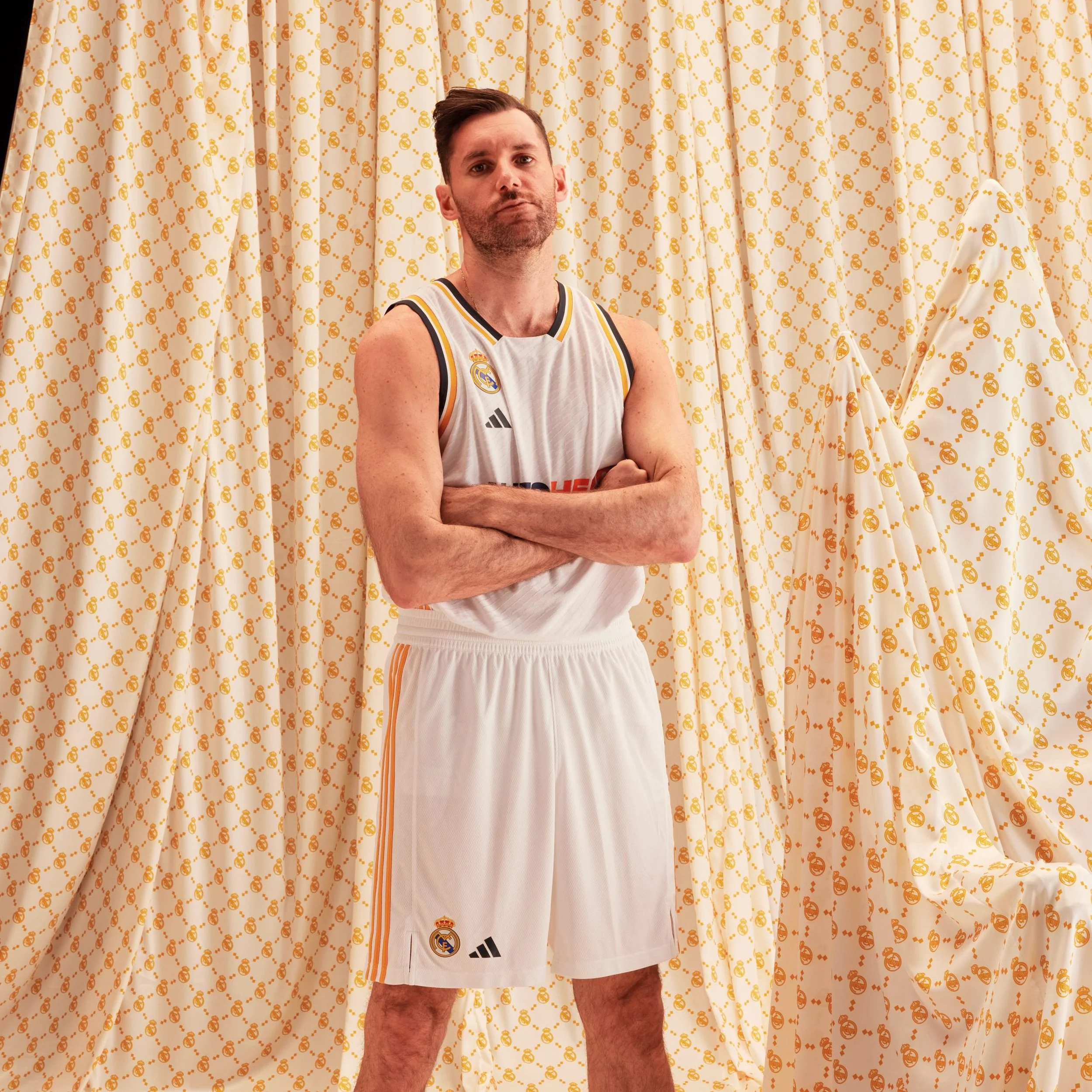 Un hombre con uniforme de baloncesto de adidas Real Madrid posa con los brazos cruzados frente a cortinas blancas decoradas con pequeños logotipos dorados de la marca
