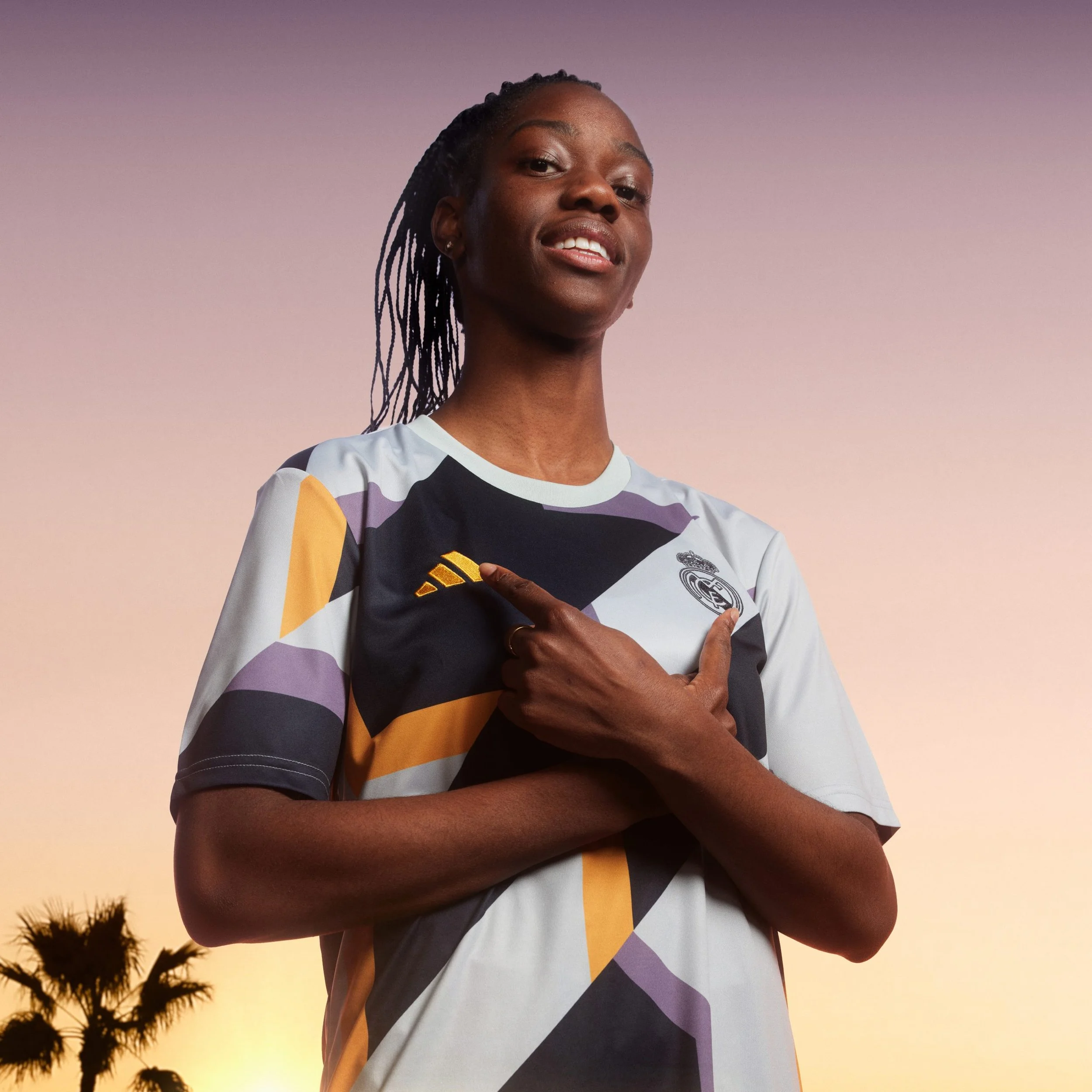 Mujer con camiseta de fútbol del Real Madrid, señalando su escudo con una sonrisa, al atardecer con palmeras en el fondo.