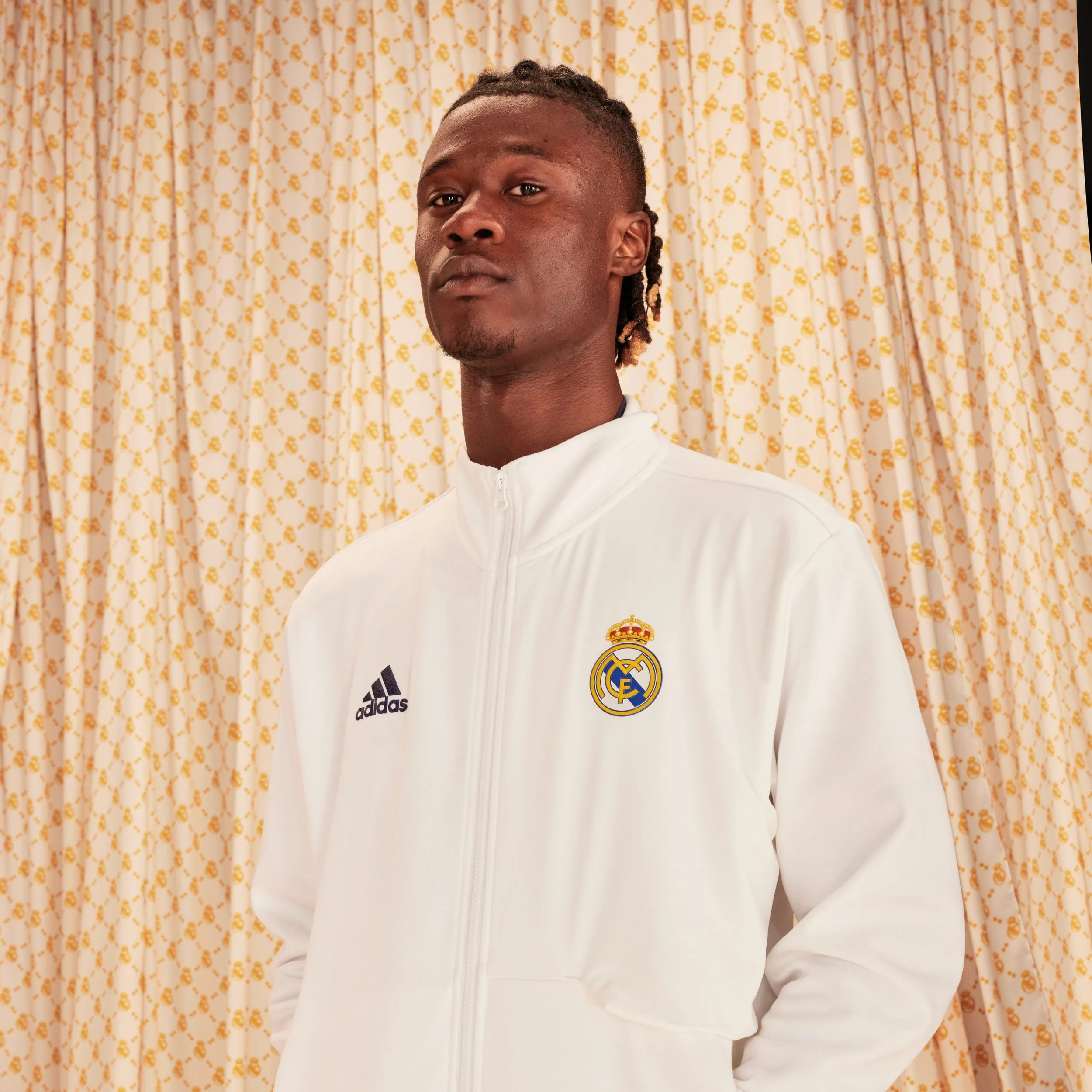 Joven con camiseta deportiva blanca adidas con el logotipo de Real Madrid, posando frente a cortinas con estampado en tonos beige y dorado.