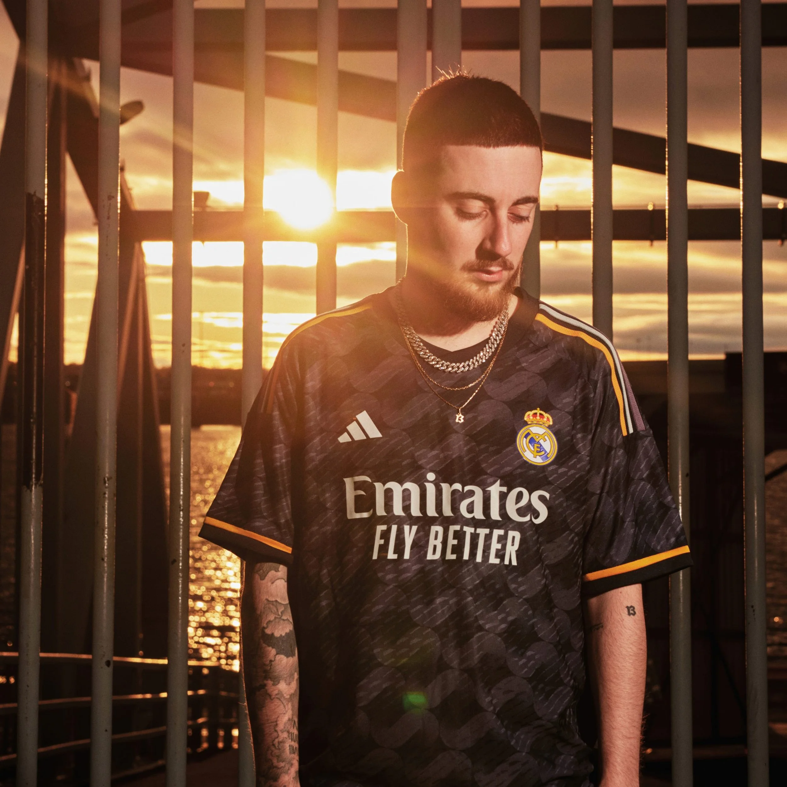Joven con barba y tatuajes, con camiseta del Real Madrid, de pie en un paseo junto al agua durante el atardecer, con barras metálicas de fondo y el sol en el horizonte.
