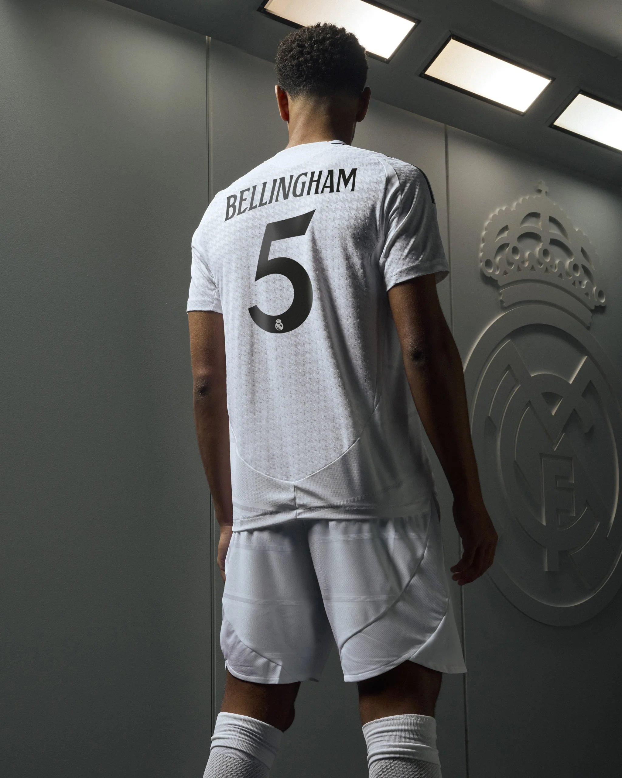 Jugador de fútbol con camiseta blanca, vista desde atrás, con el nombre 'Bellingham' y el número 5, en un vestuario del Real Madrid, con el escudo del equipo en la pared a su lado.