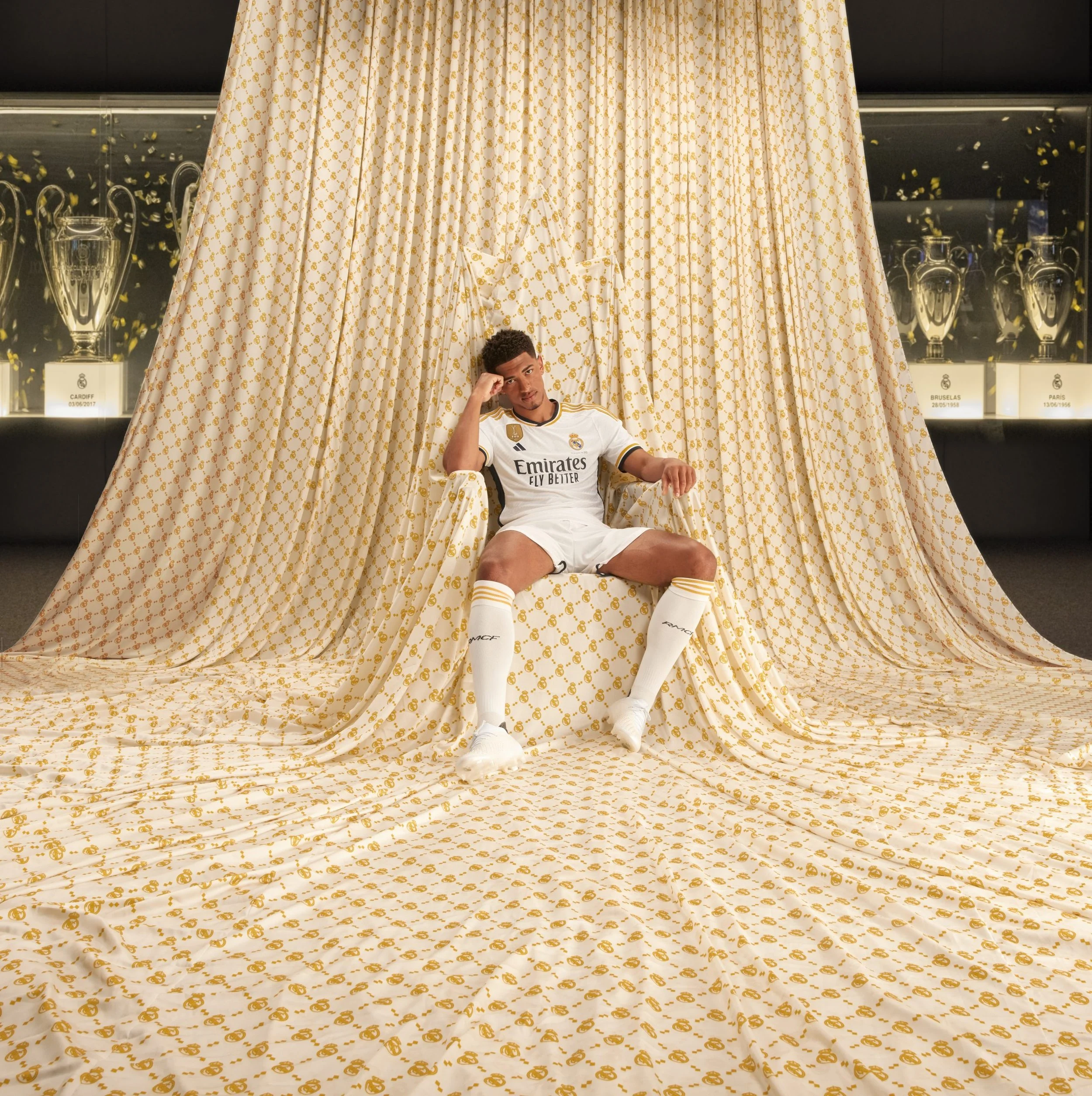 Un futbolista sentado en una silla en medio de cortinas doradas y con fondo de trofeos, vestido con uniforme de adidas Real Madrid en campaña amor incondicional