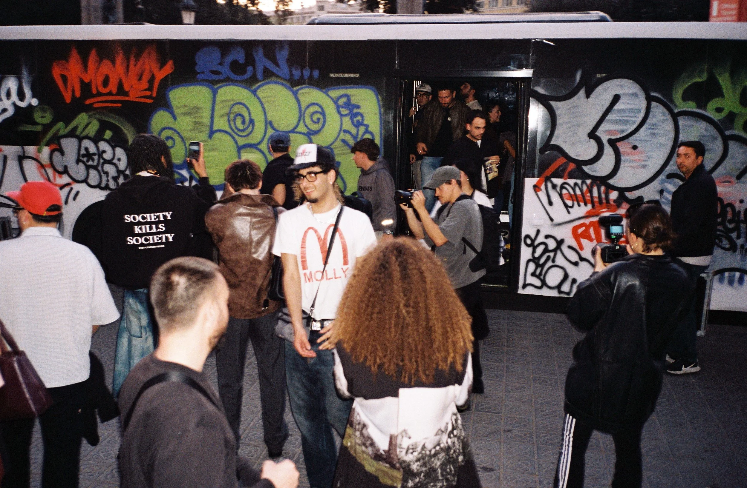 Apertura local Legit Barcelona. Grupo de personas esperando para abordar un autobús con graffiti en las paredes, algunos toman fotos y otros conversan.