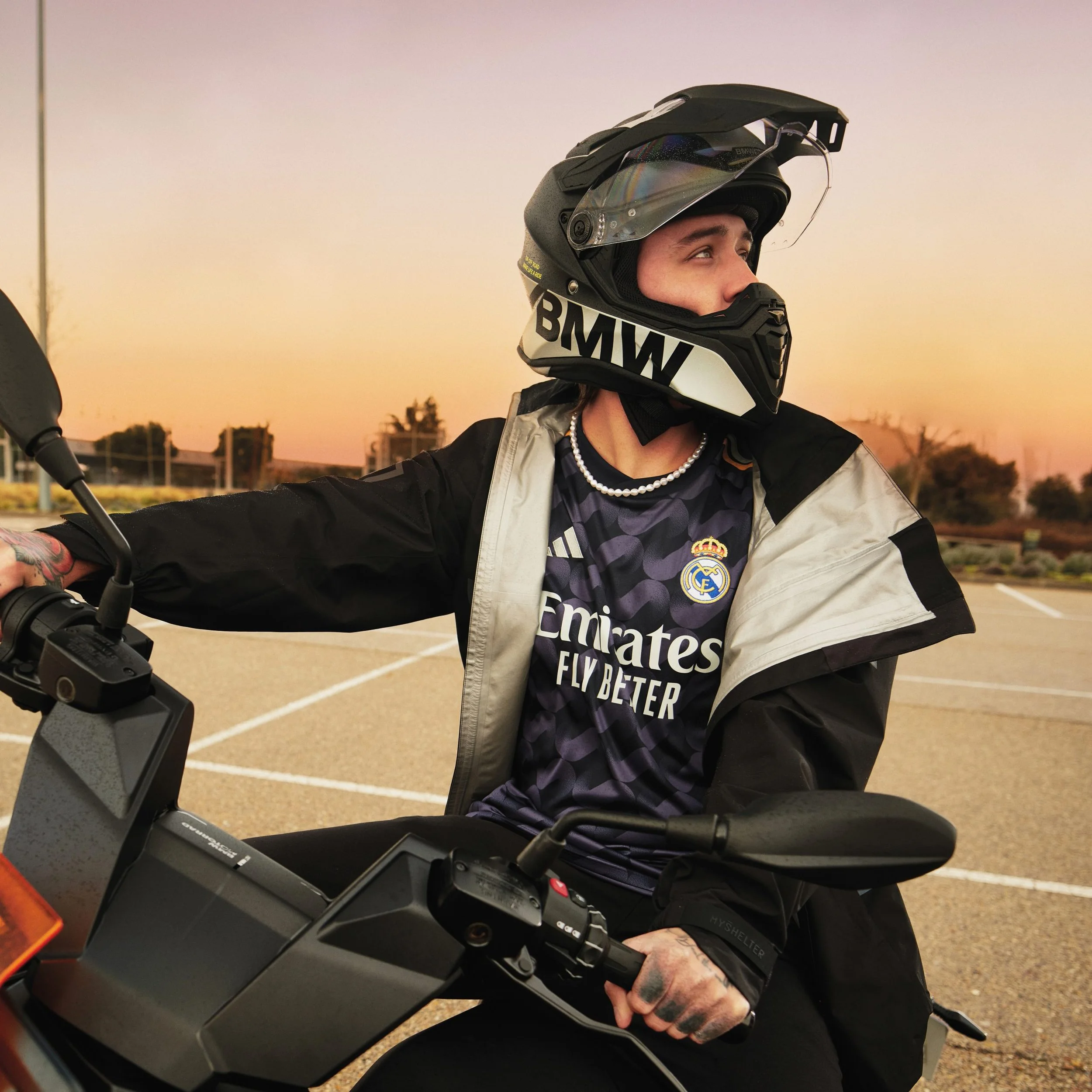Persona con casco de motociclista sentado en una motocicleta, usando una camiseta del Real Madrid en un estacionamiento al atardecer.