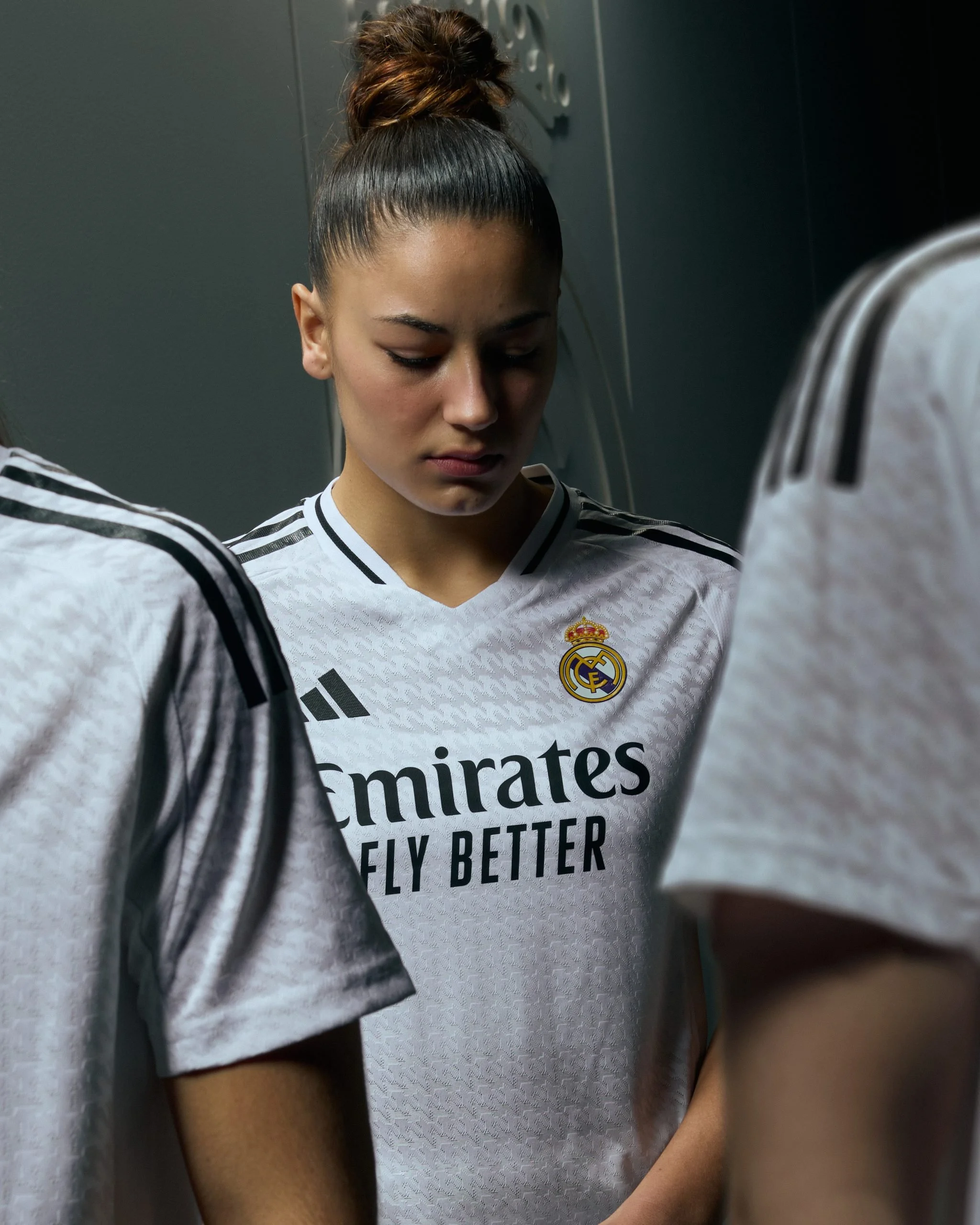 Joven futbolista en vestuario de Real Madrid, vestida de uniforme adidas, con cara pensativa y serena, en un ambiente de vestuario con otras personas a su alrededor.