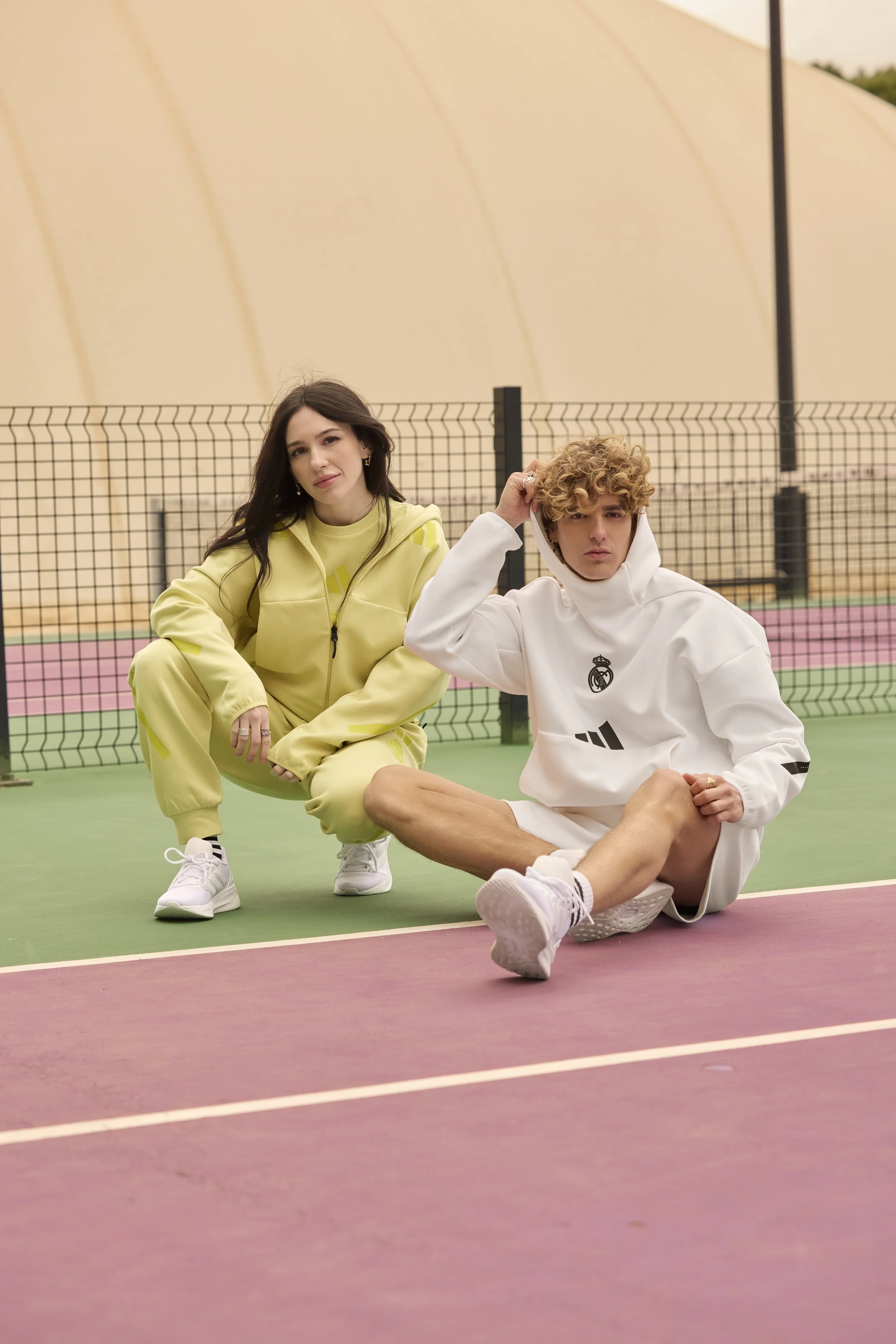 Dos jóvenes en ropa deportiva en una cancha de tenis, uno sentado en el suelo con capucha blanquiazul y la otra de pie, de color amarillo, en un entorno de cancha de color verde y rosa con fondo de estructura curva beige en campaña ZNE Urban x adidas