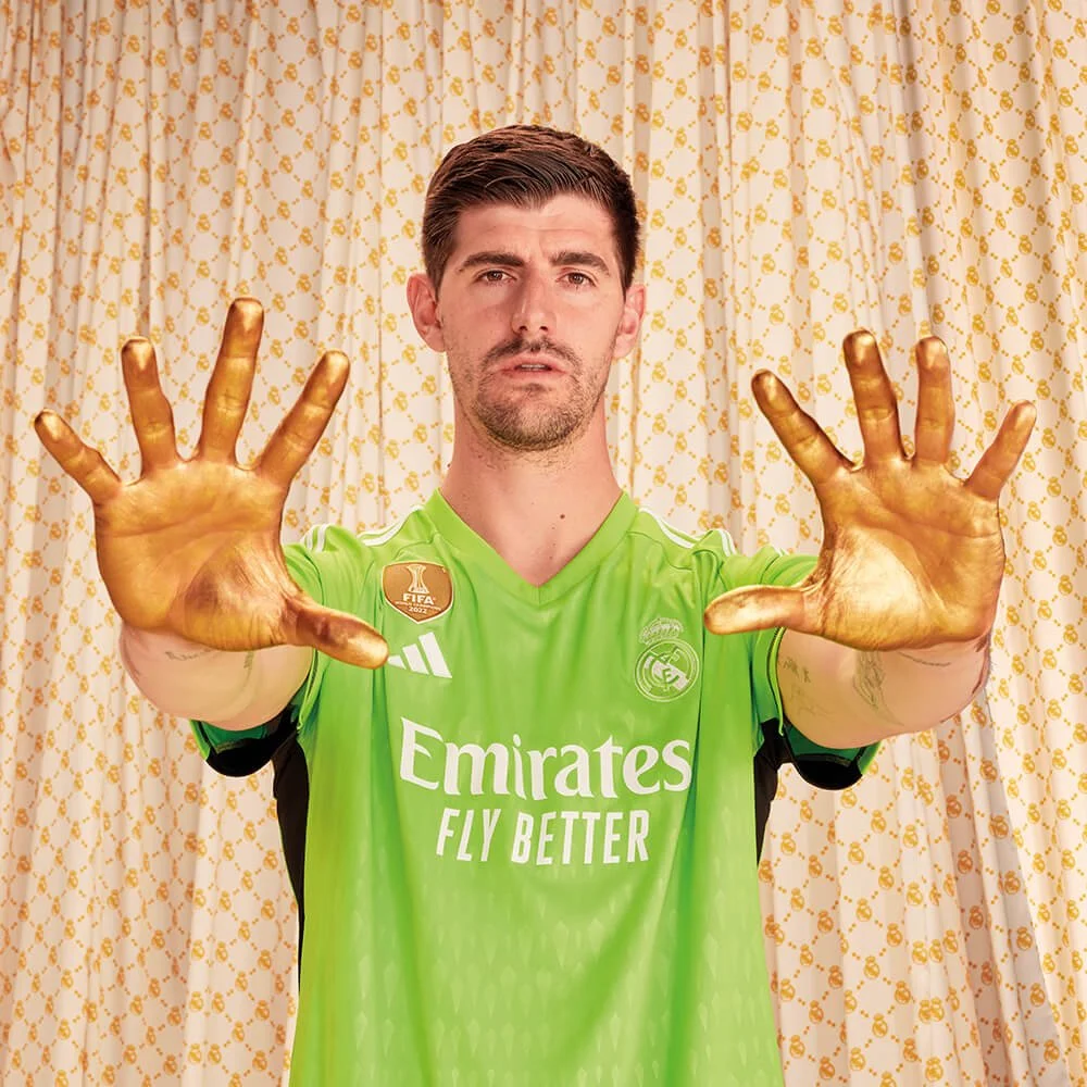 Un hombre con camiseta de fútbol en color verde de adidas Real Madrid y manos pintadas de dorado, posando frente a cortinas blancas con detalles dorados.