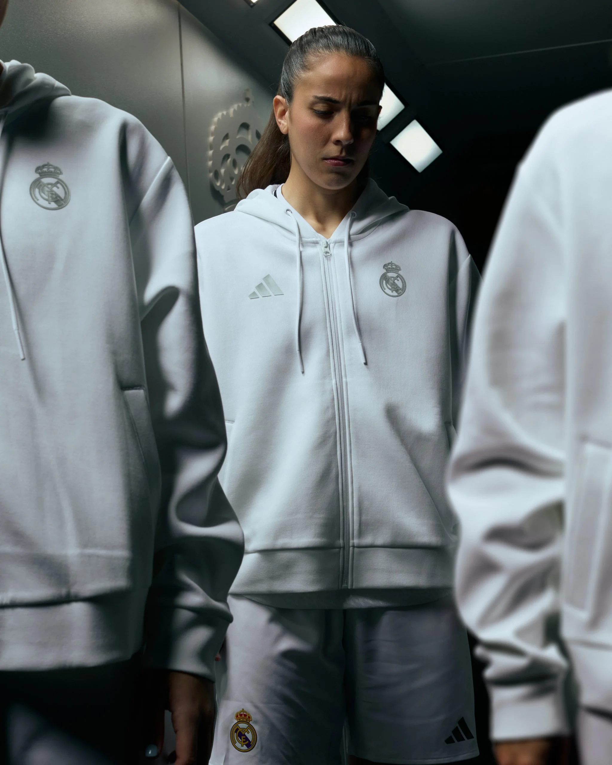 Joven deportista con ropa deportiva blanca de la Liga adidas Real Madrid, en un vestuario moderno, parece estar en un momento de reflexión o concentración.