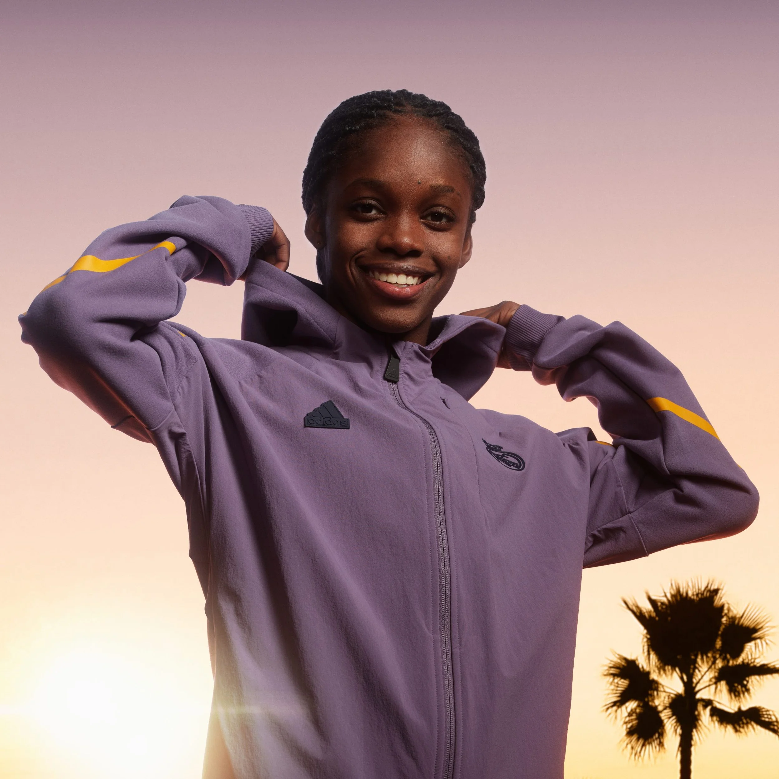 Mujer sonriendo con sudadera deportiva morada, al atardecer, con palmera en el fondo.
