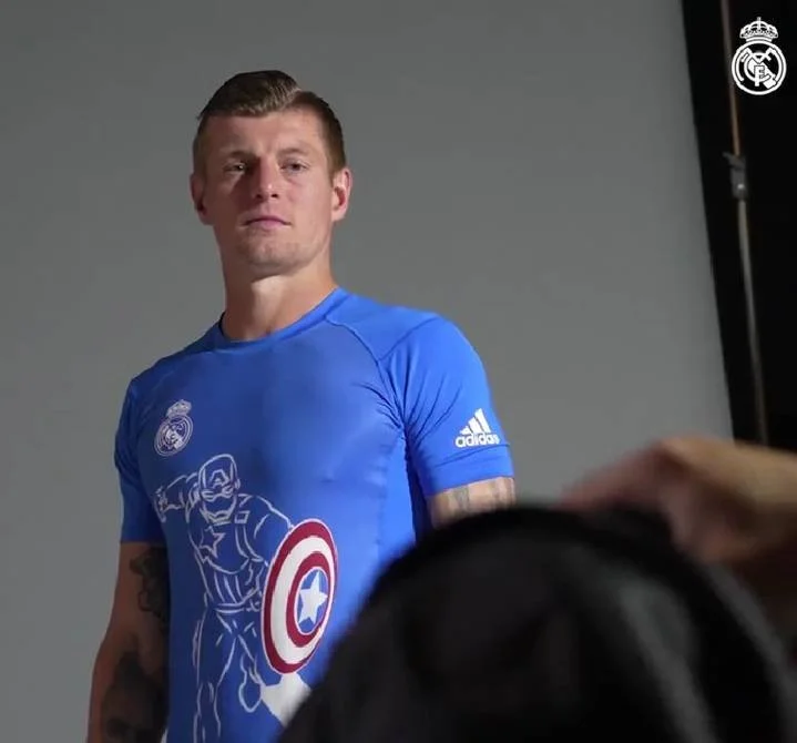 Un jugador de fútbol con camiseta azul de Real Madrid en una sesión fotográfica, con el escudo del club visible y sin sonrisa, viendo hacia la cámara. 'Heroes Among Us' con logotipos de Adidas y Marvel