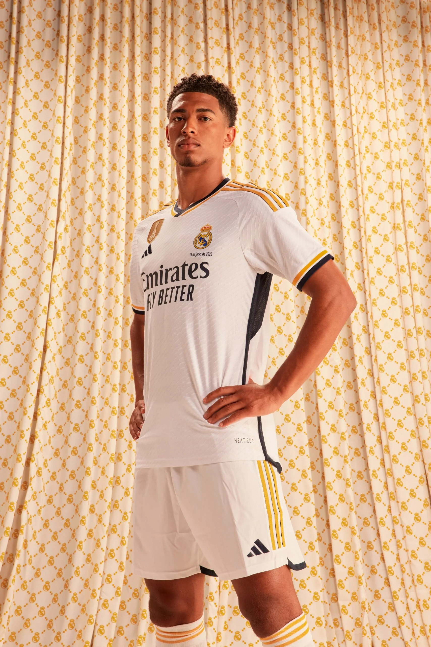 Joven futbolista con uniforme adidas Real Madrid en campaña amor incondicional en un fondo de cortinas blancas con motivos dorados, posando con las manos en la cadera.