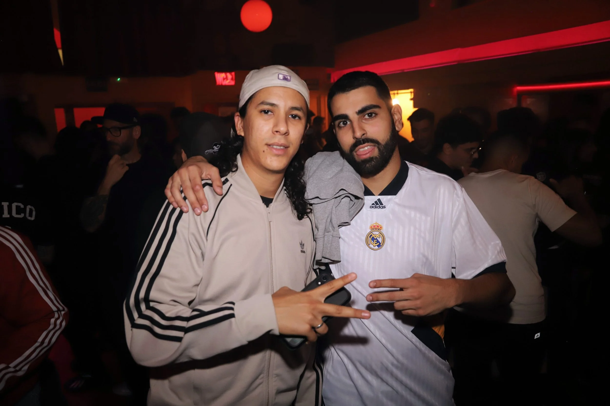 Dos hombres en evento campaña LFSTLR adidas Real Madrid, uno viste un abrigo beige con rayas negras y gorra blanca, y el otro lleva una camiseta blanca del Real Madrid, en un ambiente con poca luz y muchas personas en el fondo.