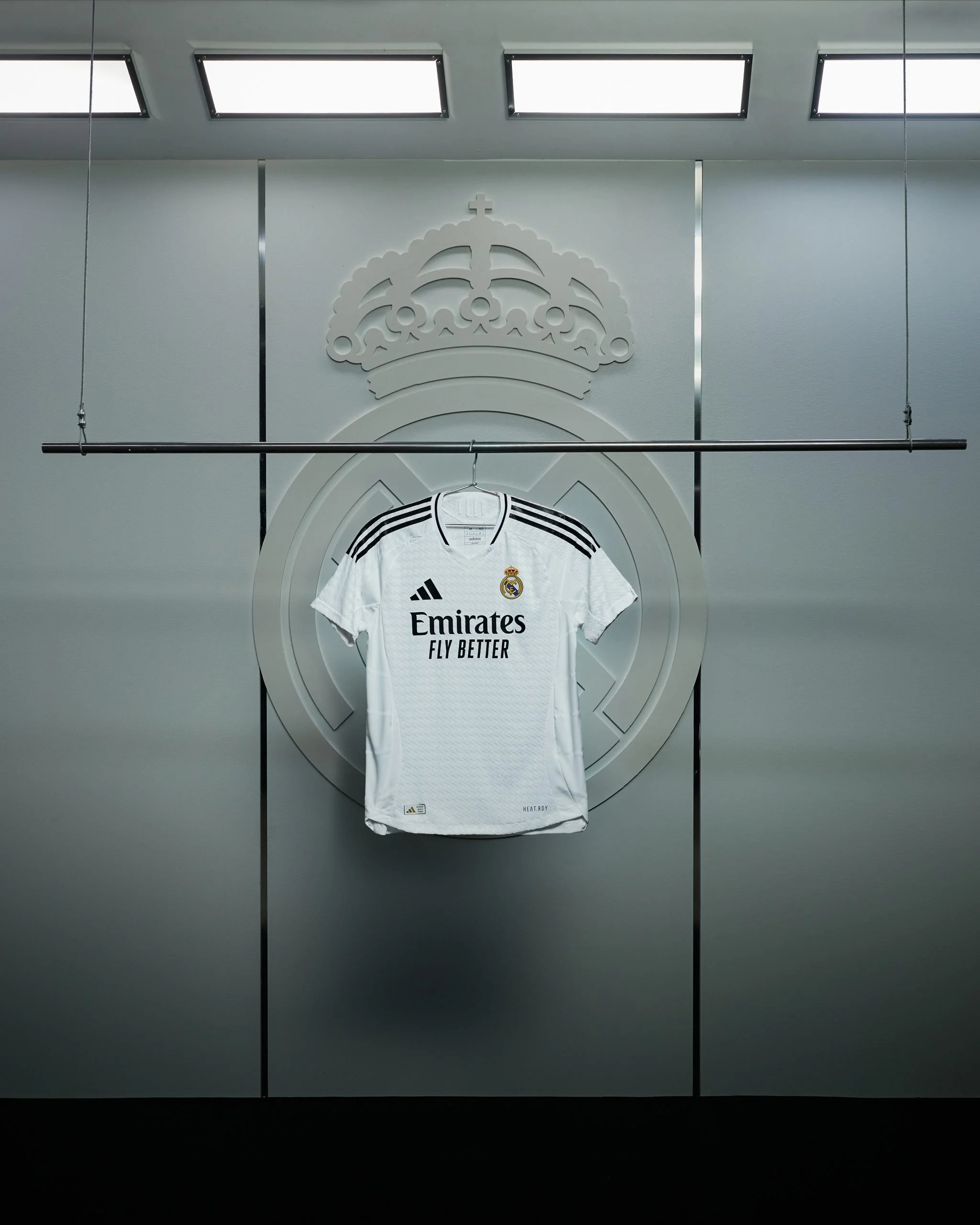 Camisa blanca adidas del Real Madrid colgada en una pared con logotipo y logo del Real Madrid en el fondo.