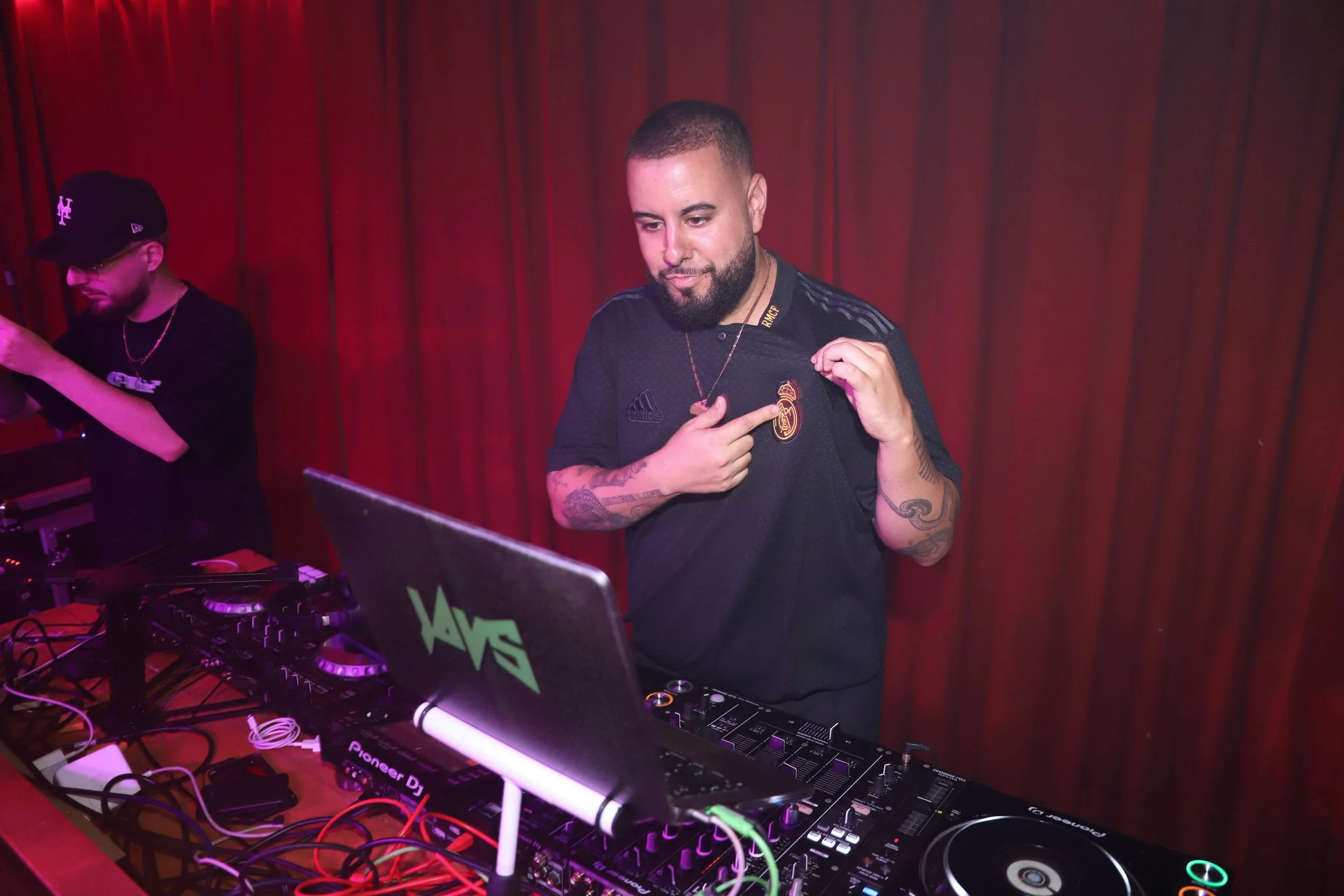 Un DJ en una cabina de música con auriculares, tocando en un club con cortinas rojas de fondo, vistiendo una camiseta de fútbol del Real Madrid y señalándose el escudo del equipo.
evento campaña LFSTLR adidas Real Madrid