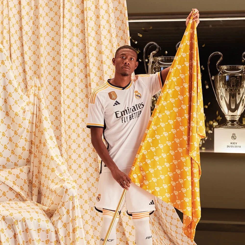 Un futbolista en uniforme de adidas Real Madrid, sosteniendo una bandera amarilla con el escudo del equipo, en un entorno decorado con cortinas a juego, con trofeos en el fondo y confeti en el aire. campaña AMOR INCONDICIONAL