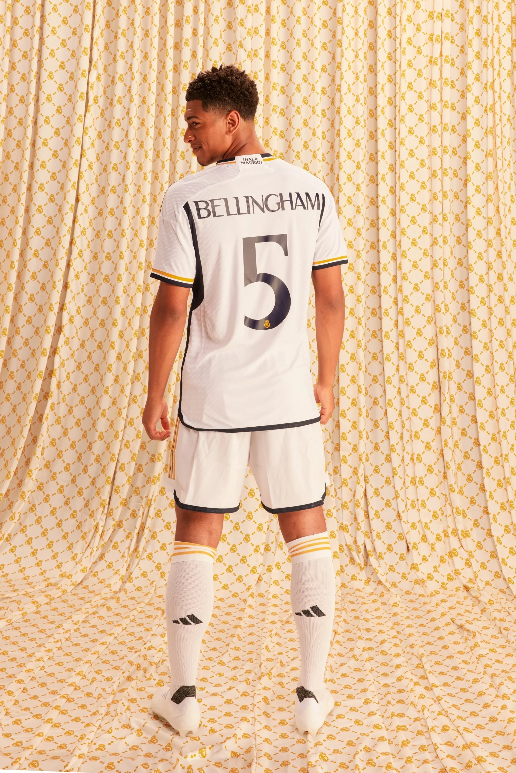 Jugador de fútbol vistiendo uniforme adidas Real Madrid en campaña amor incondicional con el número 5 y el apellido Bellingham, de espaldas, frente a cortinas doradas con logotipos del club.