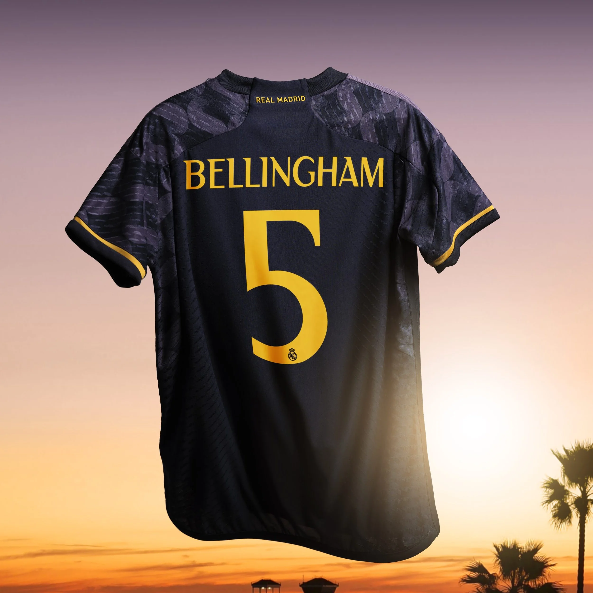 Una camiseta del Real Madrid negra con detalles en morado y amarillo, con el nombre 'Bellingham' y el número 5 en amarillo en la parte trasera, colgada contra un atardecer con palmeras y casas en el fondo.