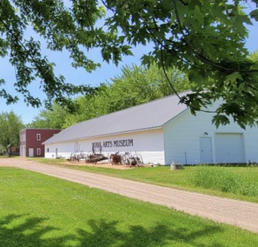 A white barn with the words 'Billy Clark's Museum' on it, a gravel path leading up to it, green grass and trees, a clear blue sky.