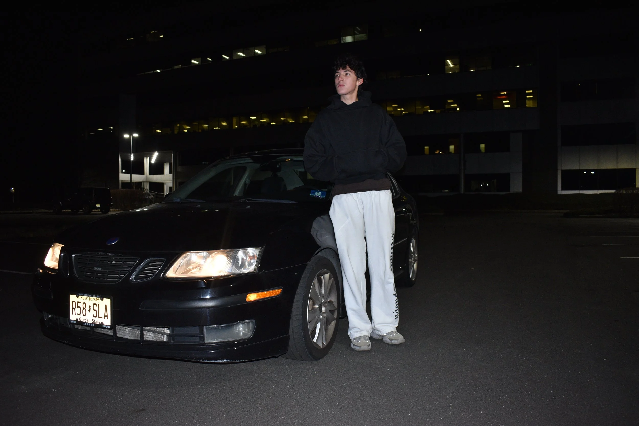 A young person in a blue hoodie and white cap standing in an empty parking lot at night, with the moon and streetlights visible in the dark sky.