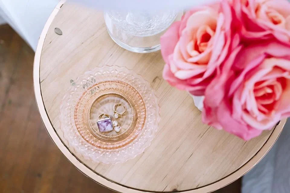 Jewelry in a pink glass dish on a wooden nightstand next to pink roses in an organized bedroom.