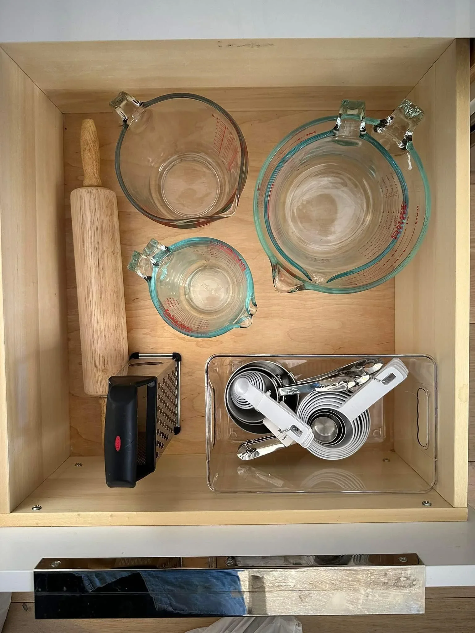 Organized kitchen drawer with glass measuring cups, a rolling pin, and a clear bin for measuring spoons.