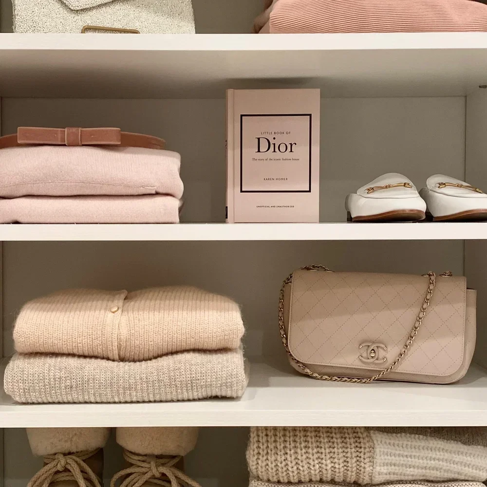 Professionally organized white closet shelves featuring neatly folded pink sweaters, white loafers, and a beige quilted bag.