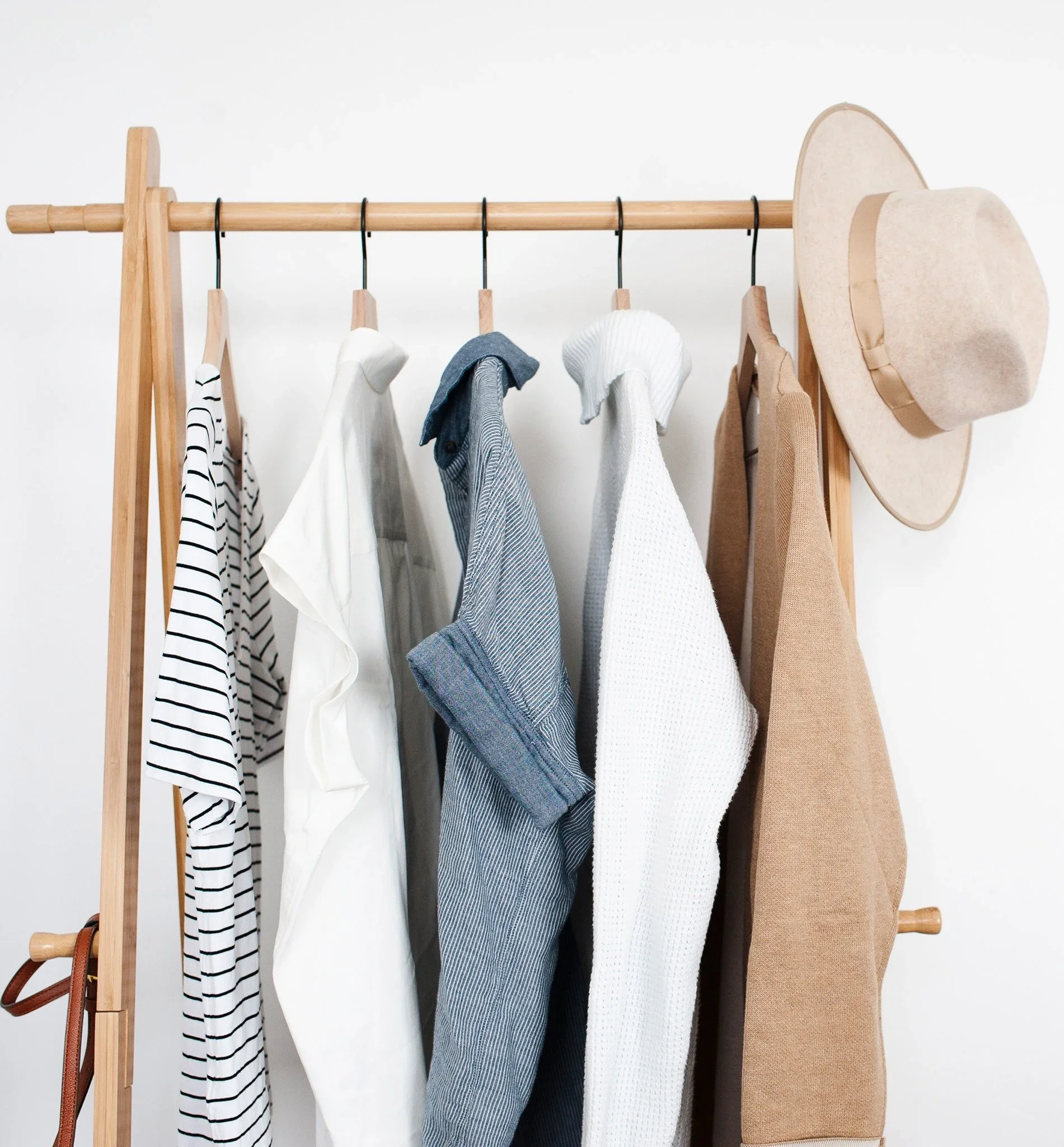 A minimalist capsule wardrobe on a wooden clothing rack with neutral-toned clothing and a tan hat, organized by Clear Spaces in Minneapolis.