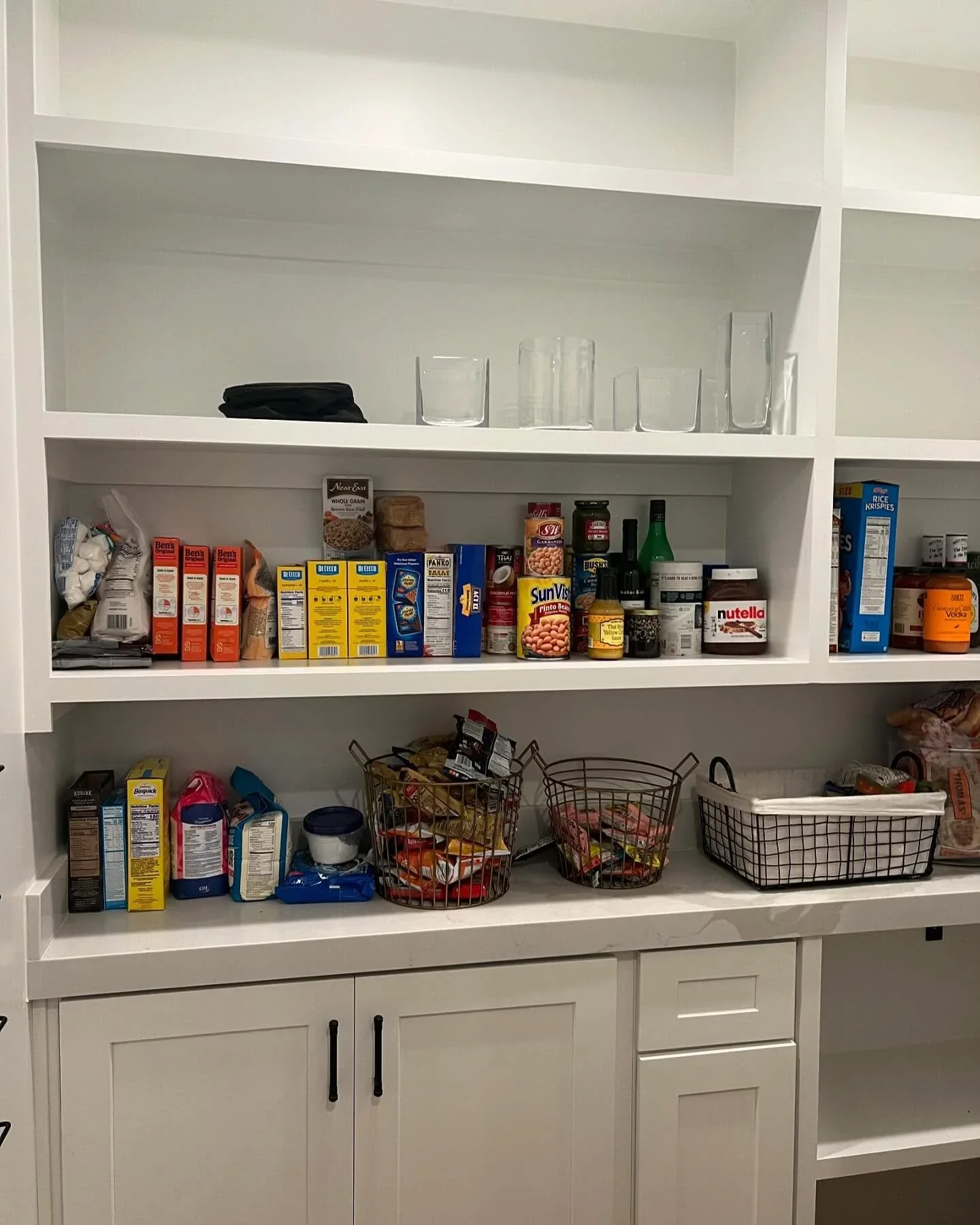 A cluttered white pantry with unorganized food boxes, jars, and wire baskets on deep shelves.