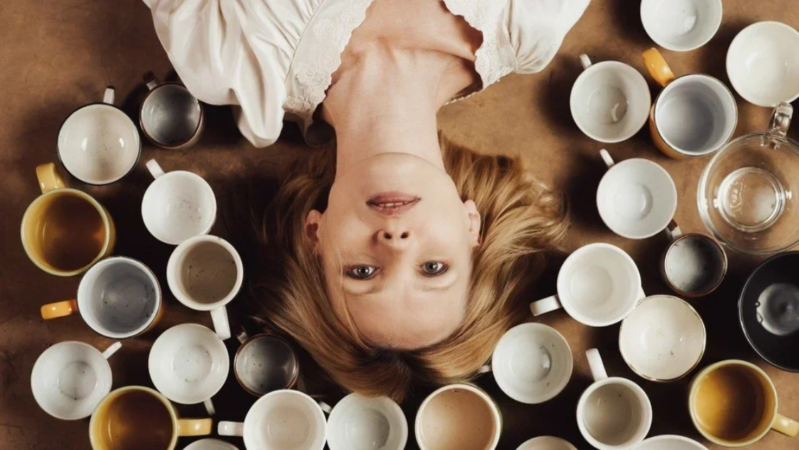 A woman lying on her back, surrounded by a large collection of coffee mugs and tea cups, illustrating the feeling of being overwhelmed.