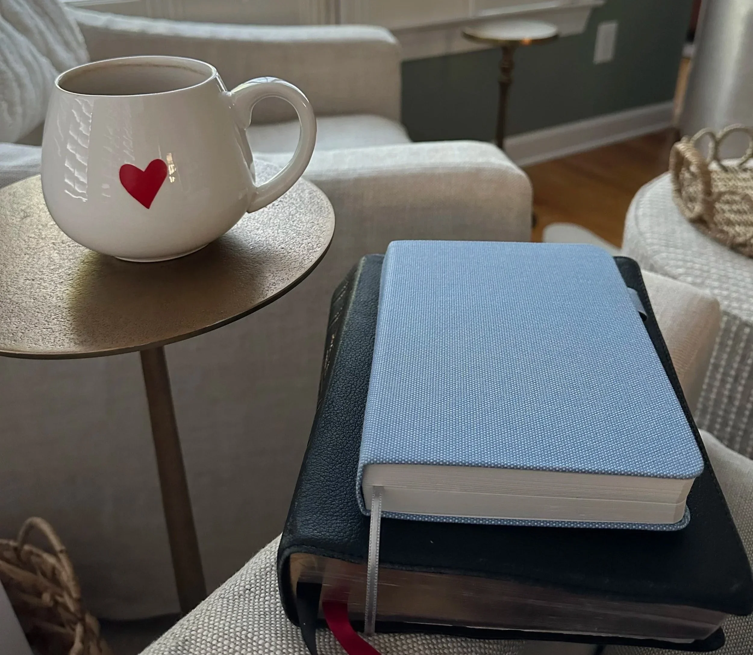 A cozy morning setup with a heart mug, a Bible, and a blue journal on a gold side table.