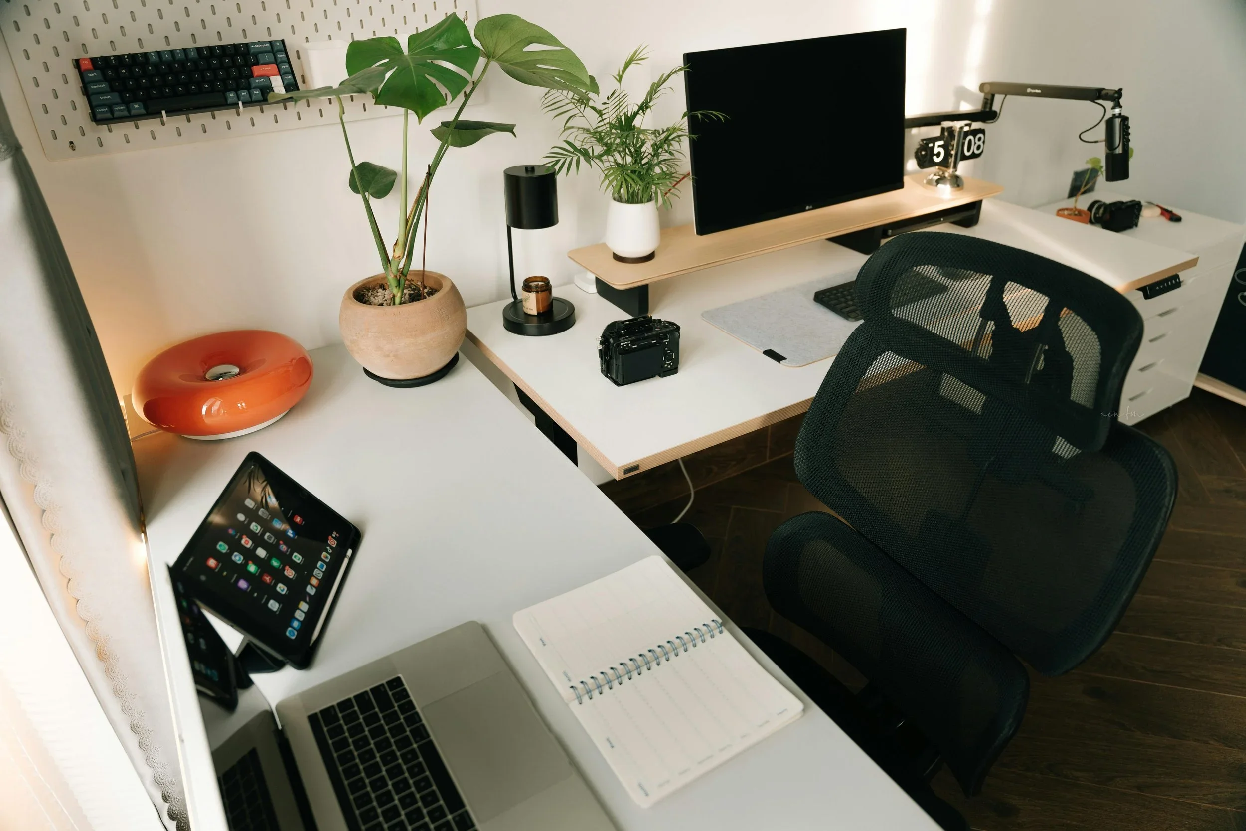 Modern organized home office with a white L-shaped desk, laptop, plants, and ergonomic black mesh chair.