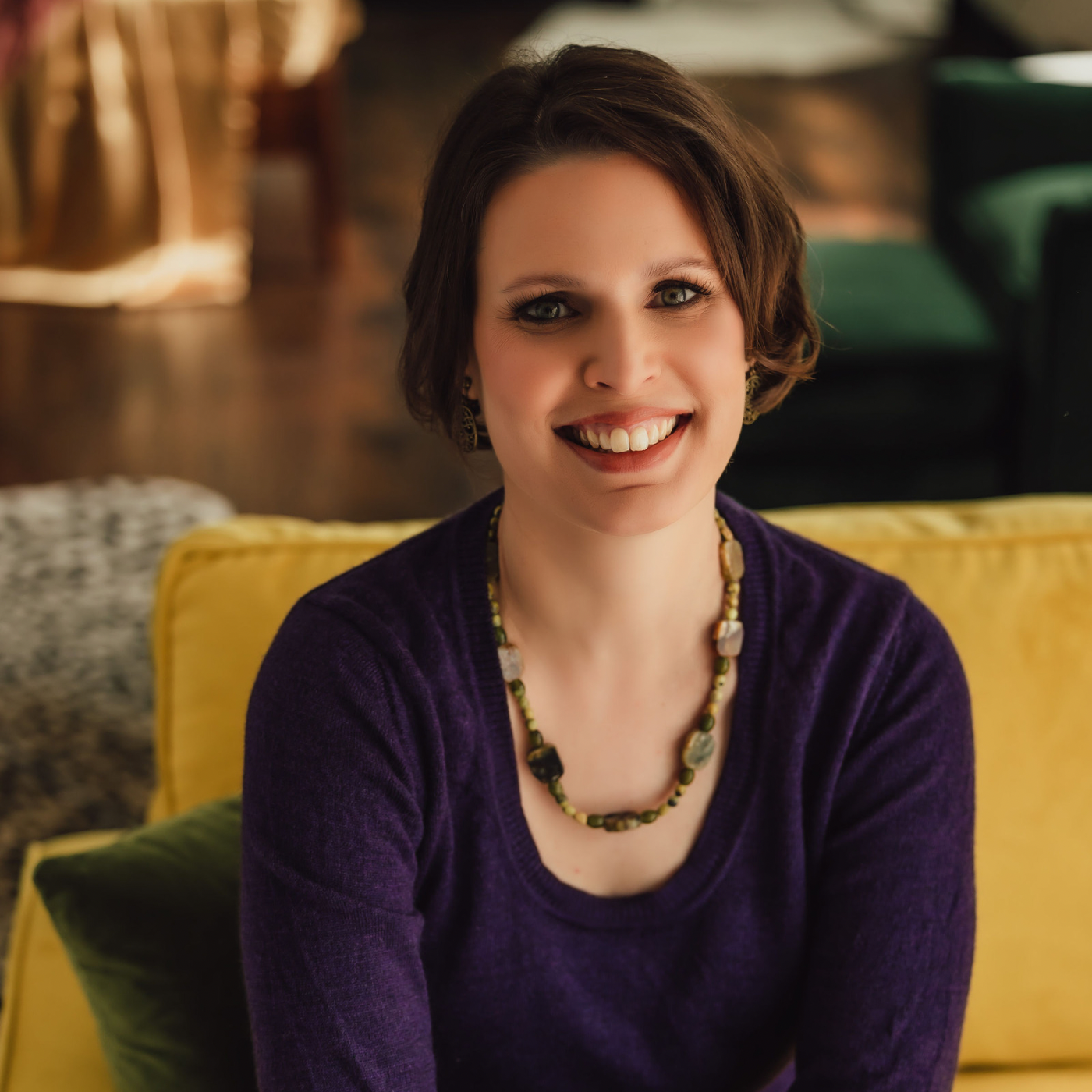 Professional organizer Kelly Weltin, founder of At Your Assistance LLC, smiling in a purple top on a yellow sofa.