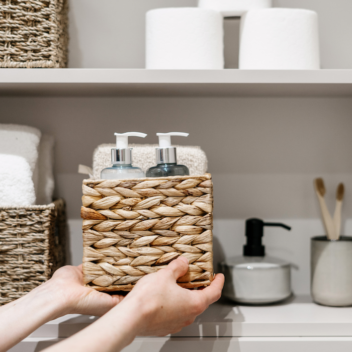 Hands placing a woven water hyacinth basket on an organized bathroom shelf.