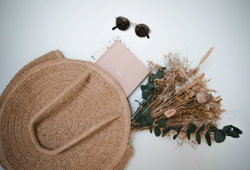 A flat lay of lifestyle essentials including a round straw bag, sunglasses, a pink notebook, and dried flowers on a white background.