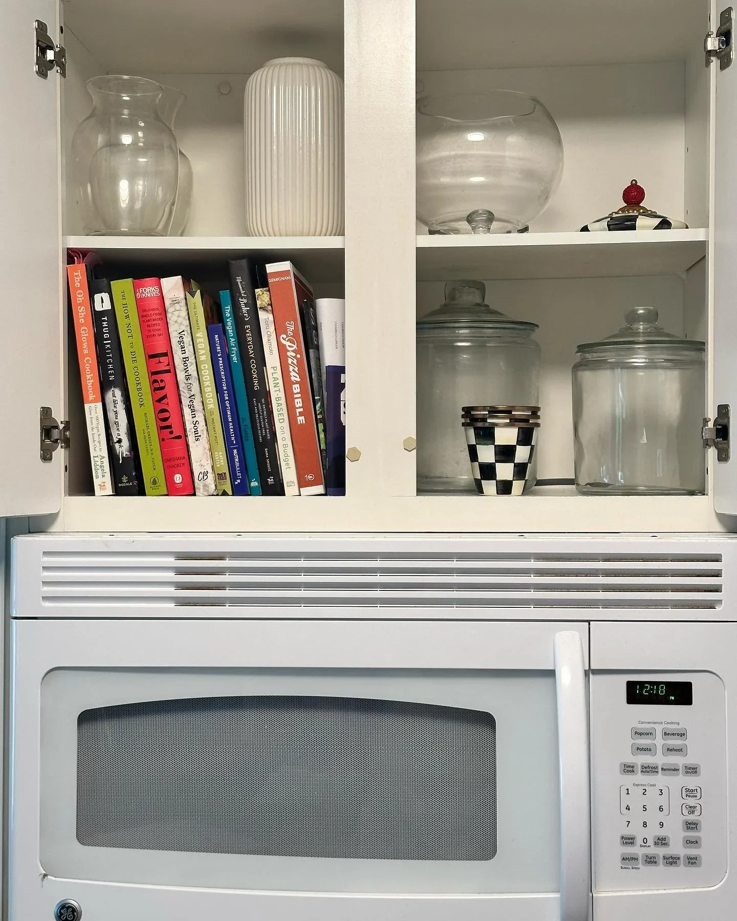 Organized white kitchen cabinet above a microwave featuring cookbooks and glass canisters by Organize to Live NC in Raleigh.