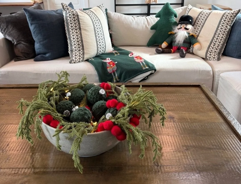 Professional holiday living room styling by Incredibly Organized in Dallas, featuring a festive bowl centerpiece with evergreen branches and a nutcracker on a white sofa.