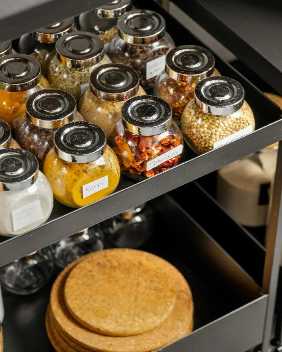 Organized kitchen spice rack with labeled glass jars and stacked wooden coasters.