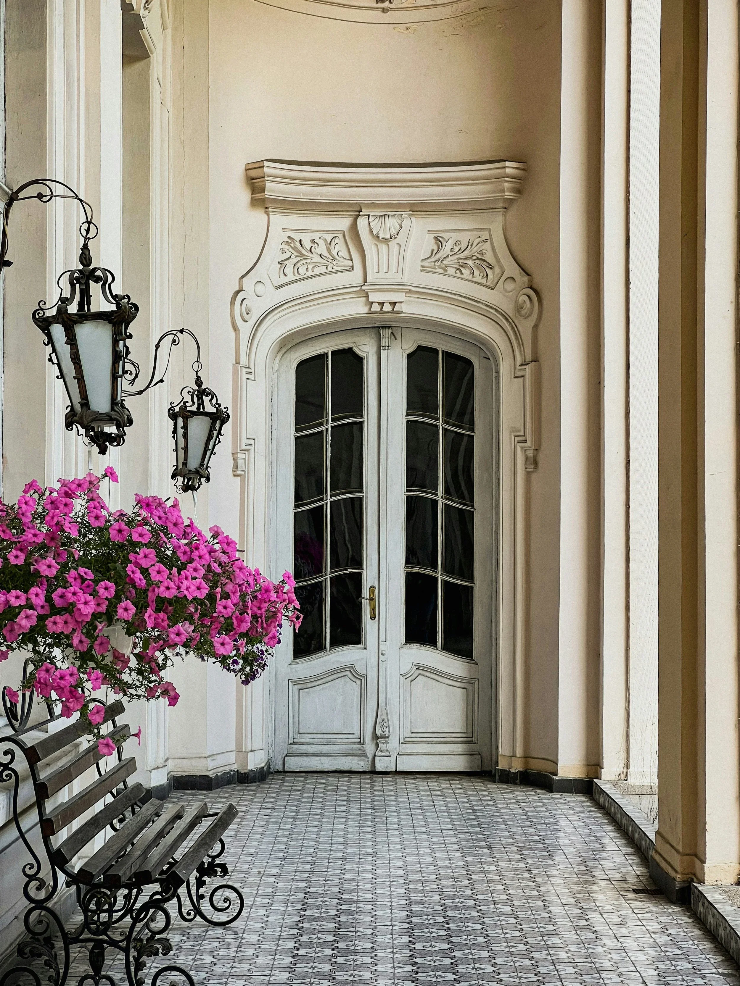 Ornate white double doors with classical stone carvings and vibrant pink flowers in a sunlit hallway.
