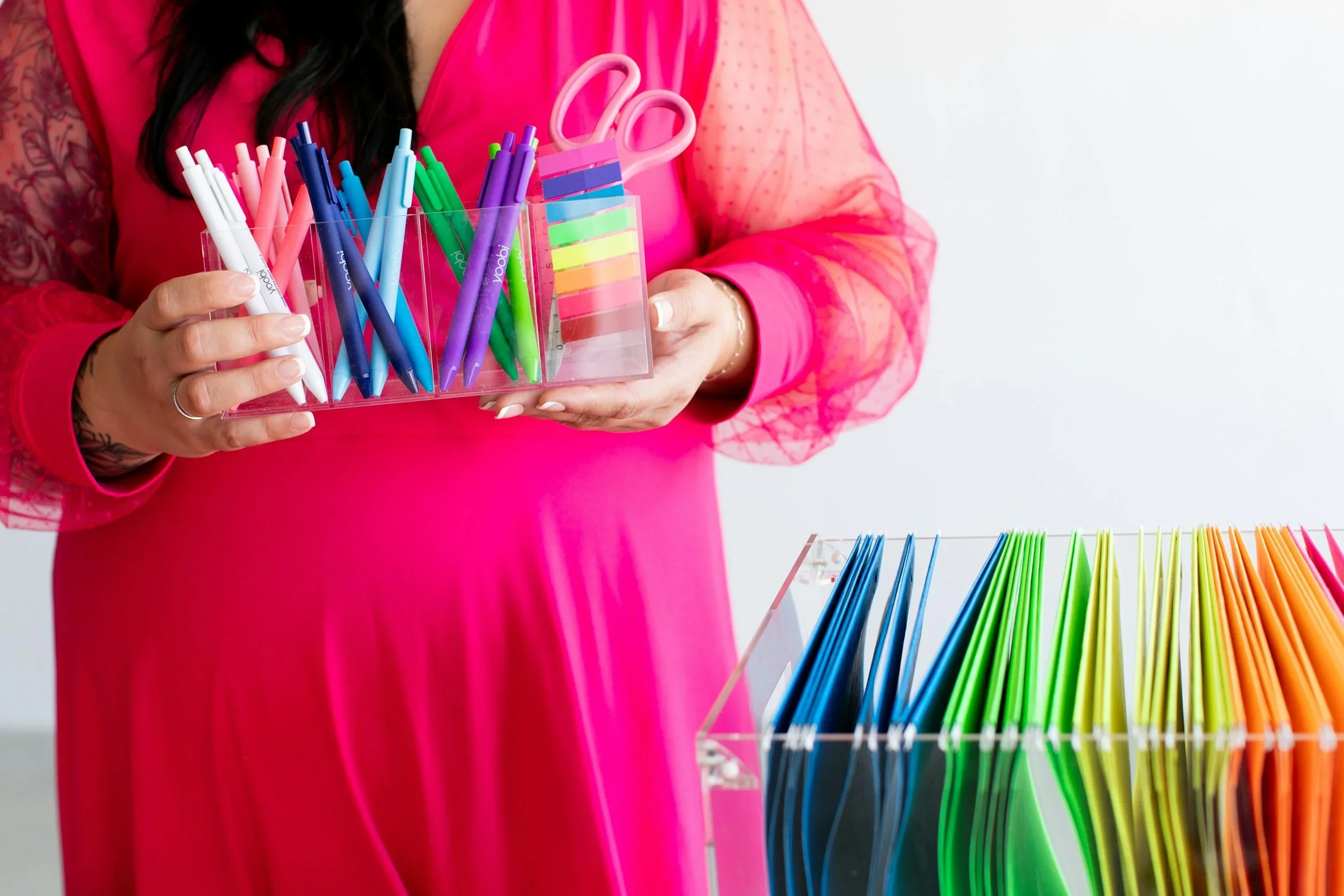 A professional organizer holding an acrylic tray of colorful pens and scissors next to rainbow hanging files.