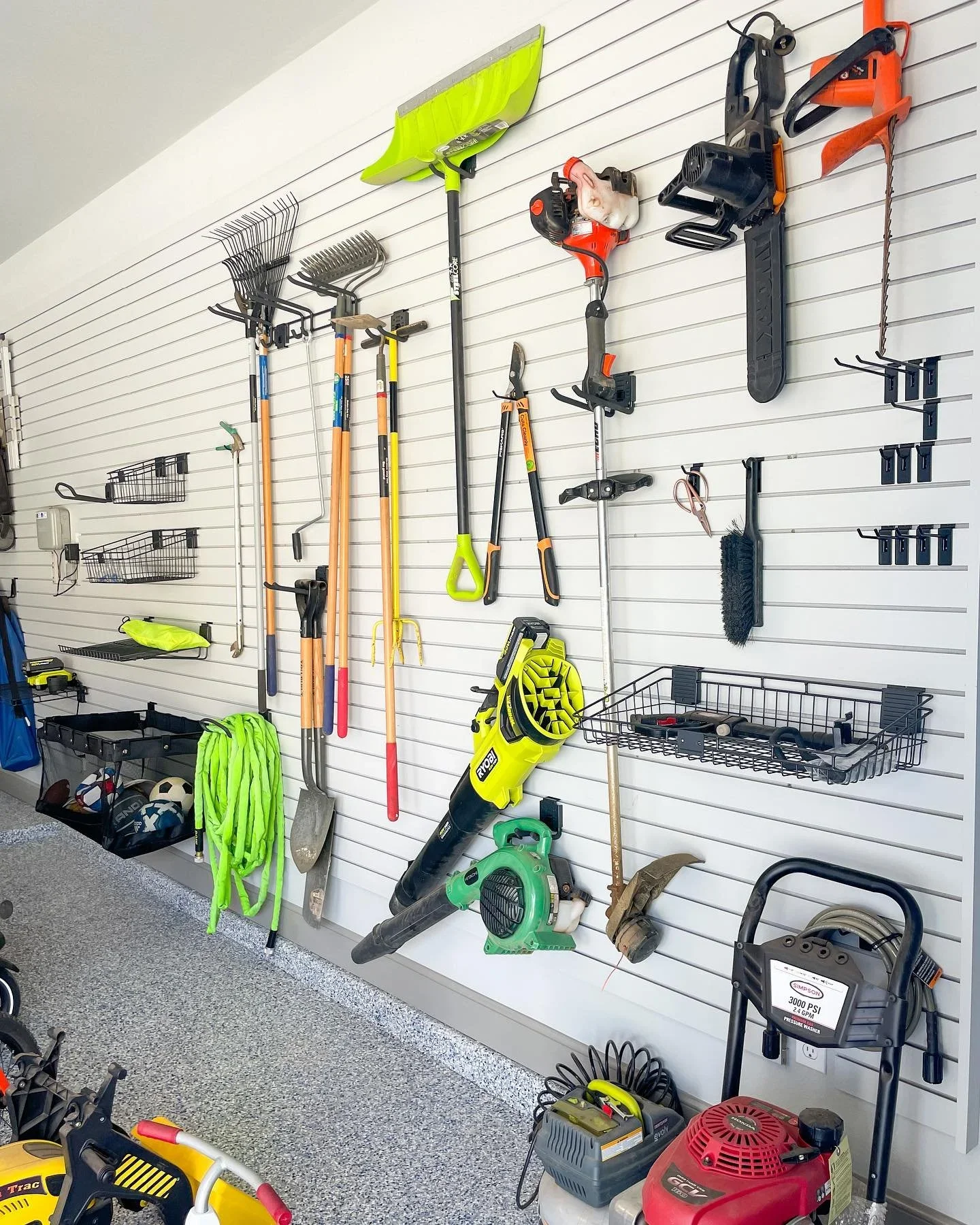 Organized garage slatwall by Minimize Then Organize featuring garden tools, leaf blowers, and sports equipment.